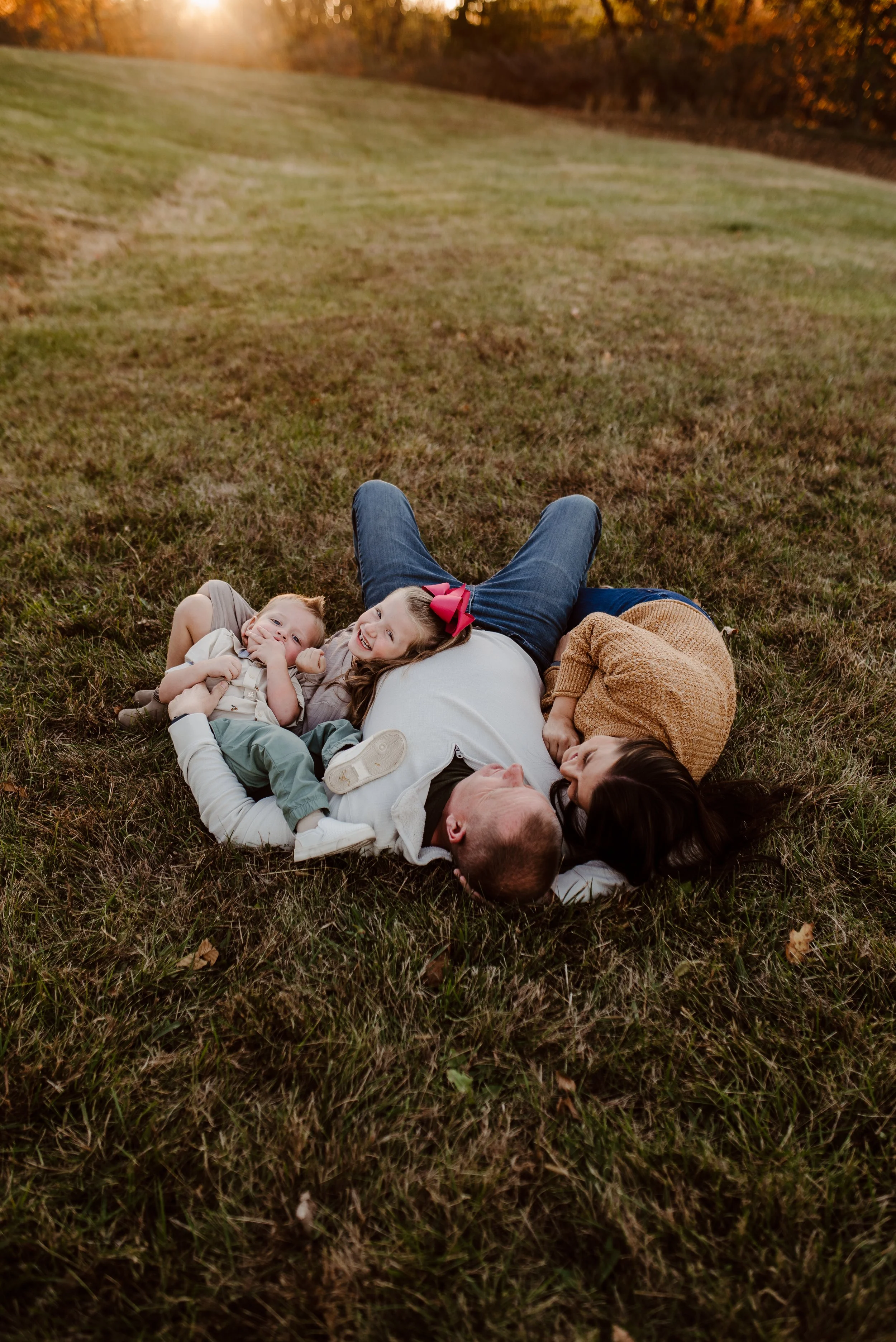A family of four lying on a grassy field during sunset, with trees in the background. The father and mother are lying face-to-face, with two children cuddled between them, smiling and enjoying a moment outdoors.