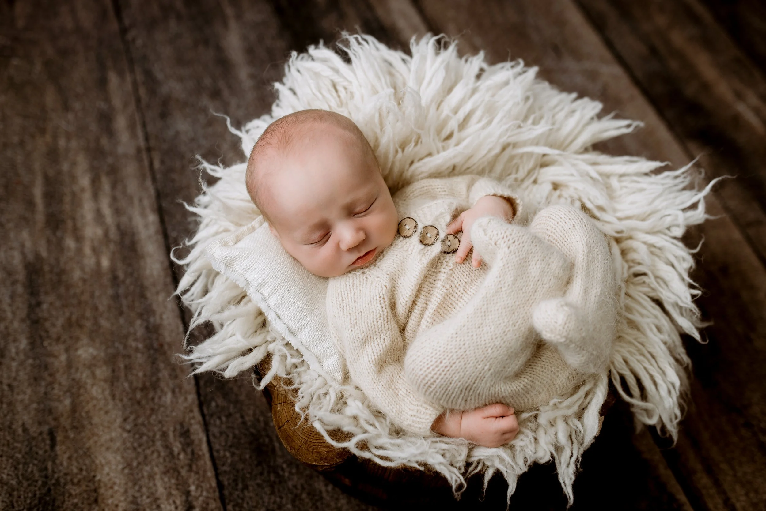 A sleeping baby dressed in a cream-colored knitted outfit, lying on a fluffy white blanket on a wooden surface.