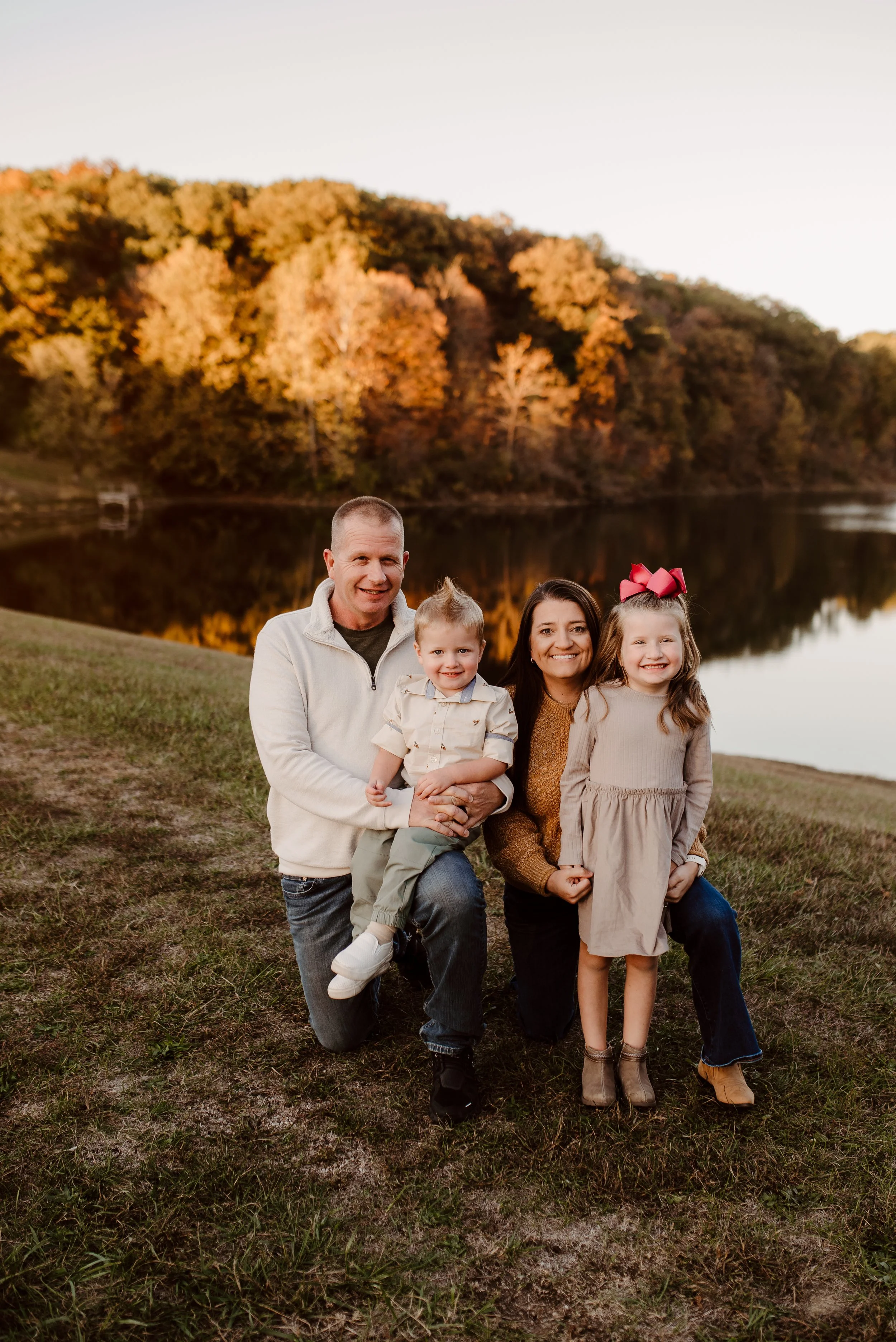 A family of four posing by a lake during autumn, with trees showing fall colors in the background. The father is kneeling on one knee holding a young boy, the mother is kneeling, and a young girl stands beside them, all smiling.