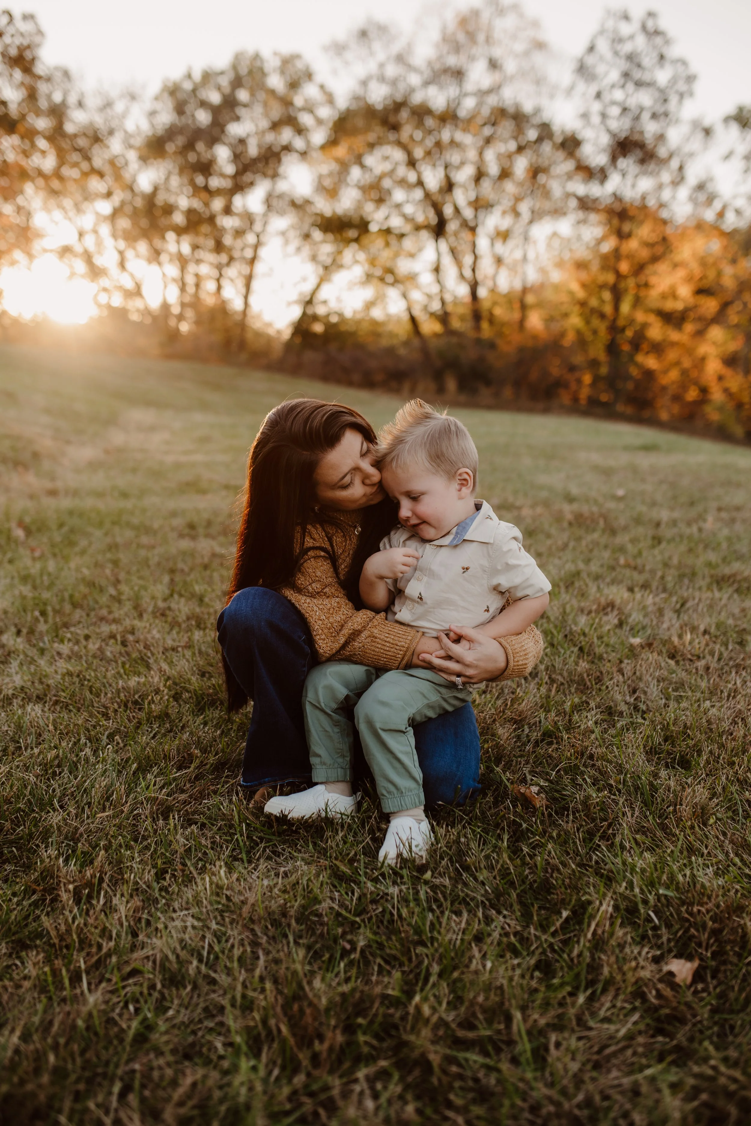 A woman and young boy sitting on a grassy field at sunset, with trees in the background, sharing a tender moment.