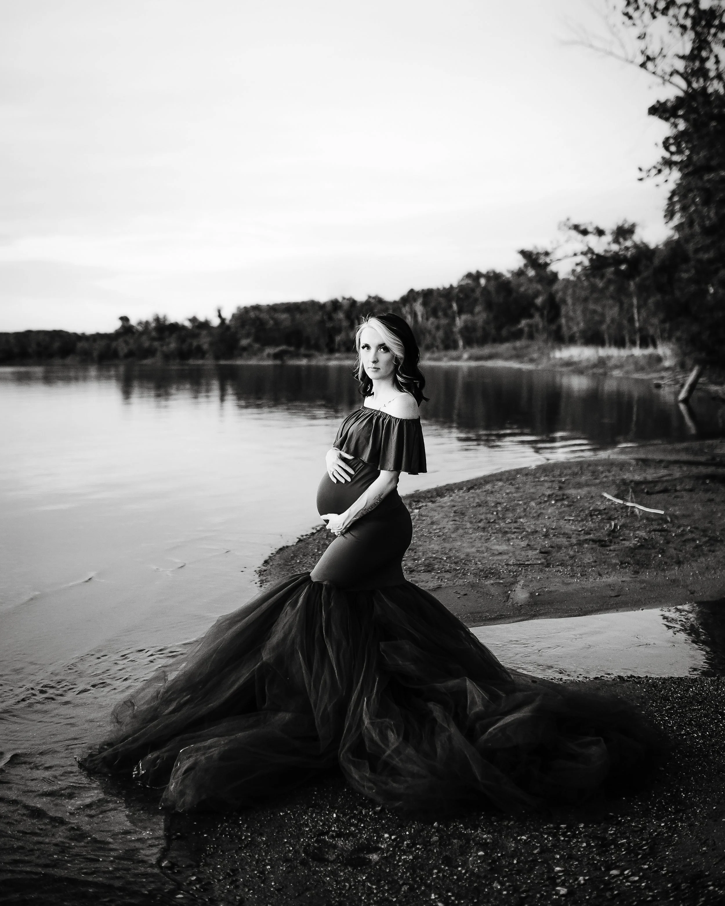 A pregnant woman in a dark tulle gown stands on a beach near a body of water, with trees in the background, in a black-and-white photograph.