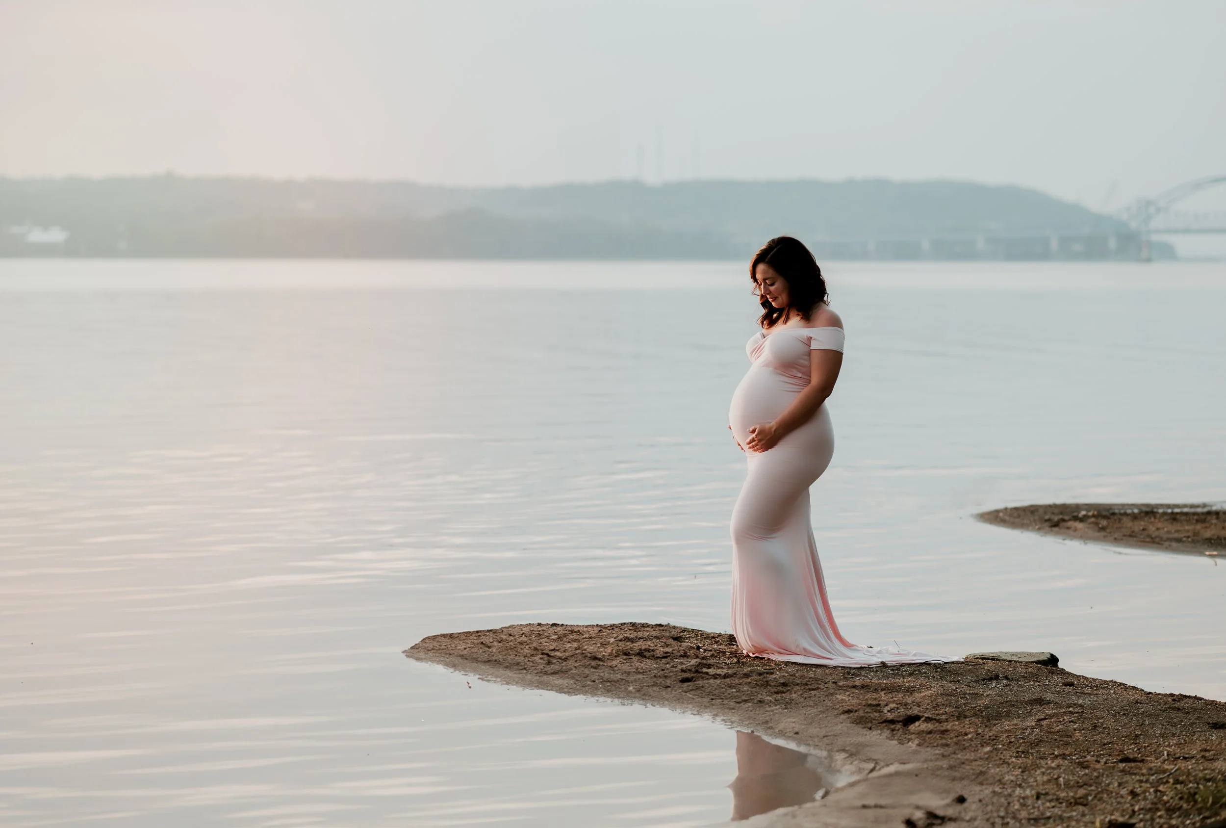 Pregnant woman in a white dress standing on a small sand and dirt peninsula by a calm body of water with a distant bridge and hilly landscape in the background.