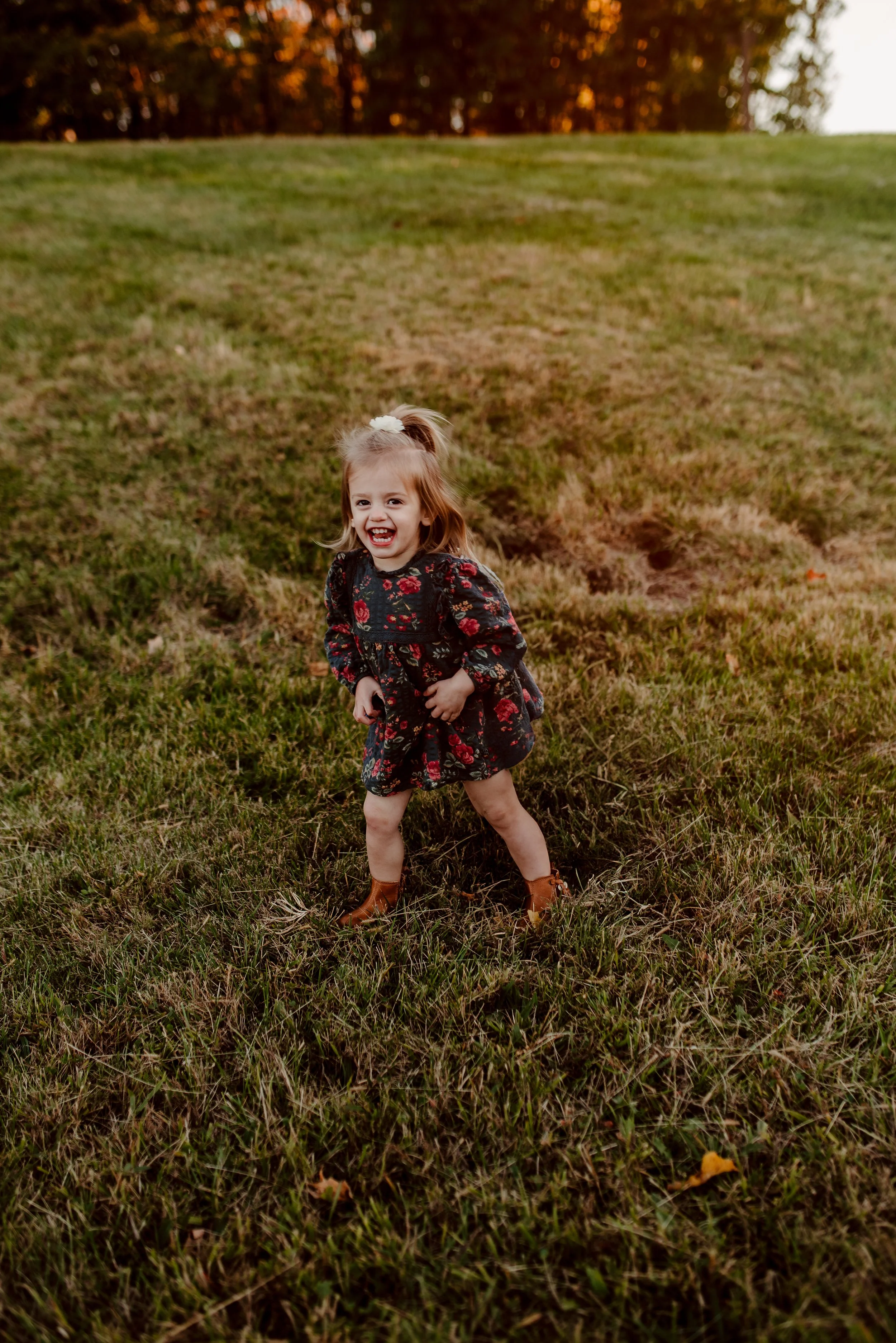 A young girl with long hair, wearing a black floral dress and brown boots, laughing and standing in a grassy field during sunset.
