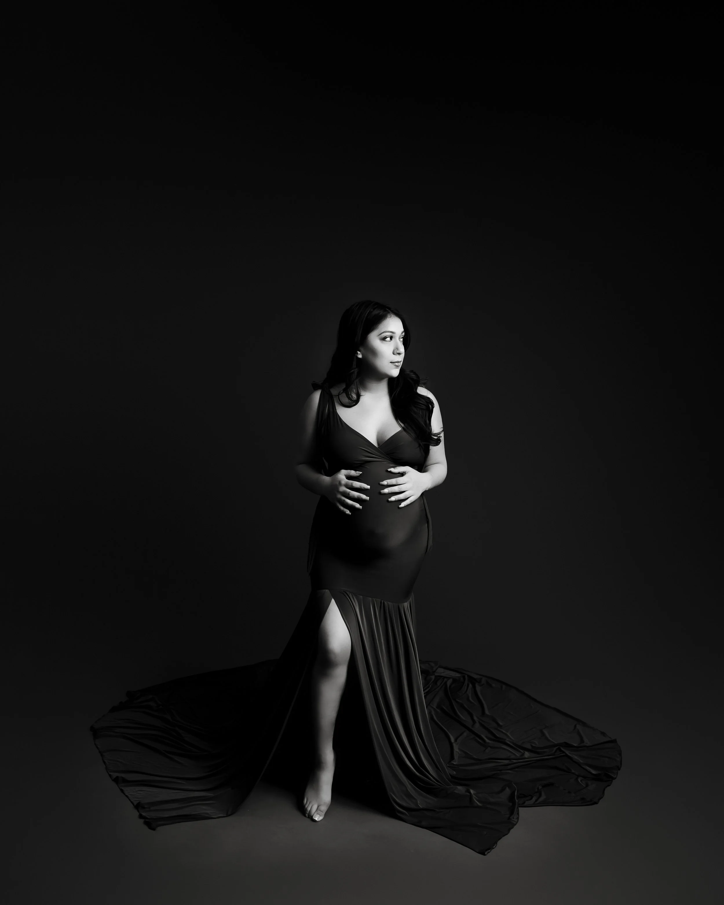 A pregnant woman in a black dress with a high slit, standing barefoot on a dark background, looking to the side.