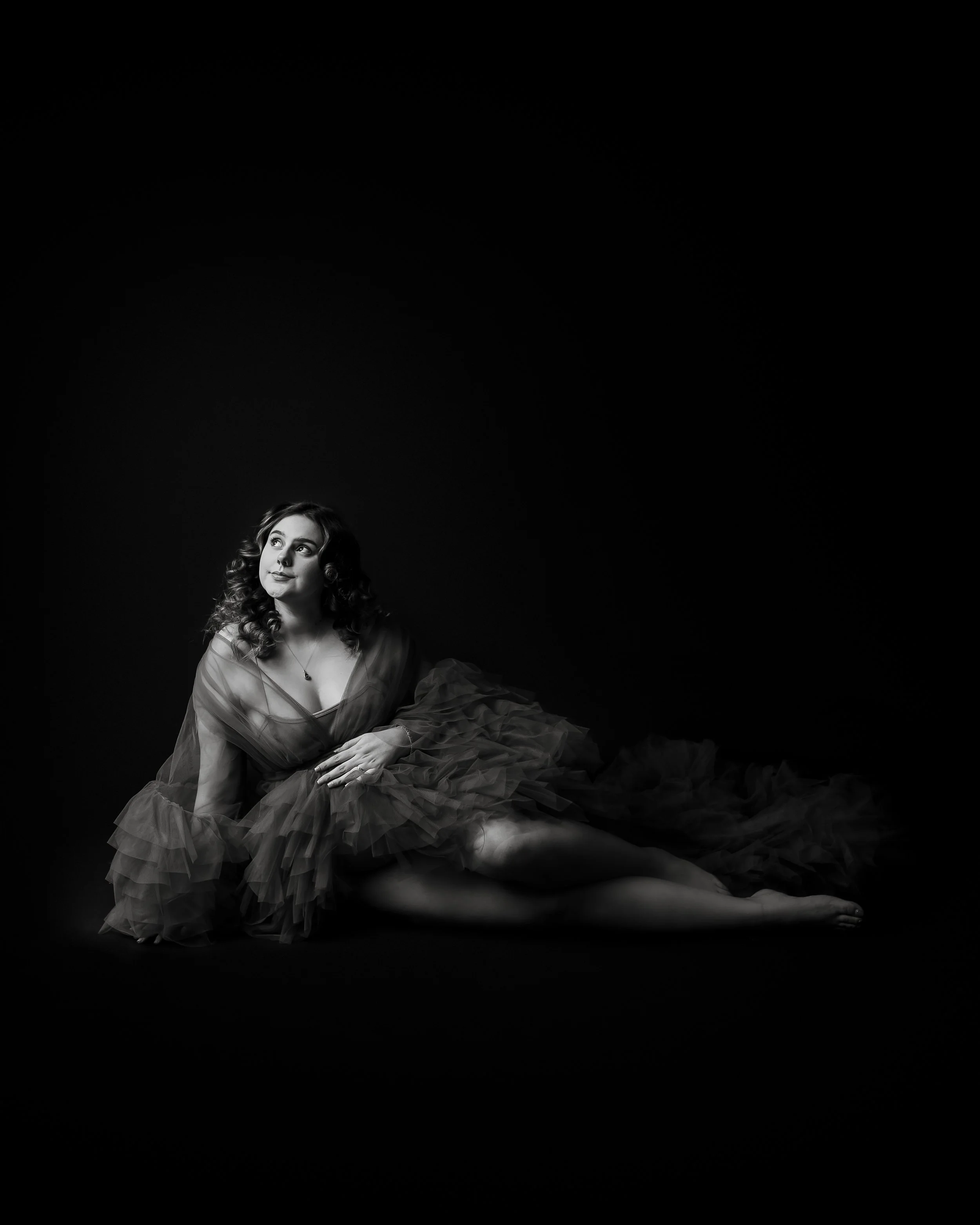 A woman with curly hair wearing a dark tulle dress, sitting on the floor against a dark background, gazing thoughtfully upward.