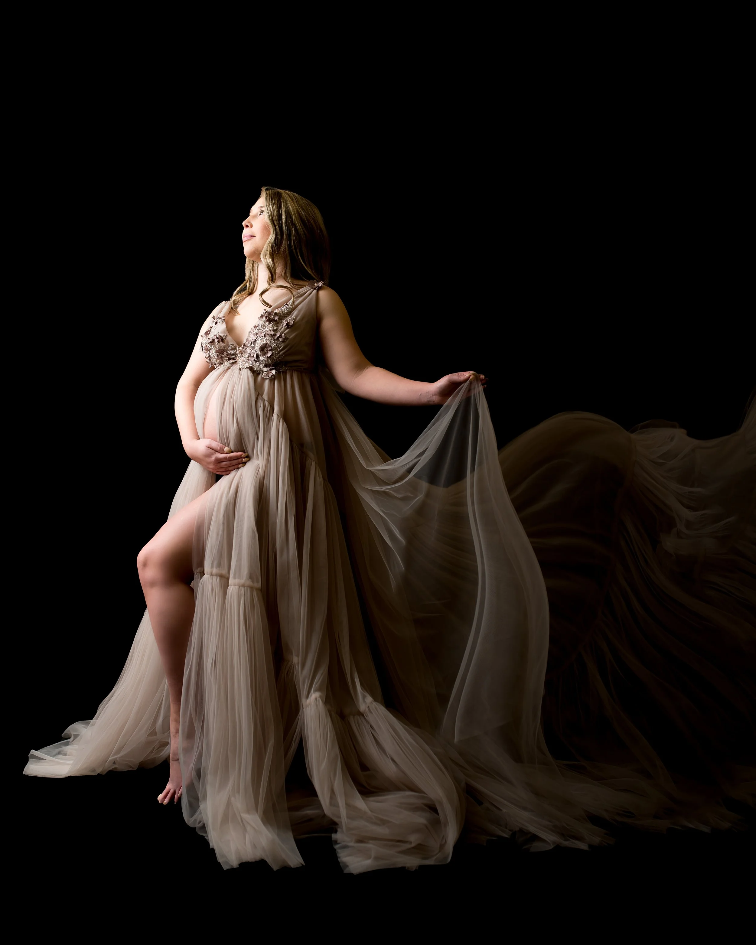 Pregnant woman in a flowing beige gown, holding her belly with one hand and lifting the gown with the other, against a black background.