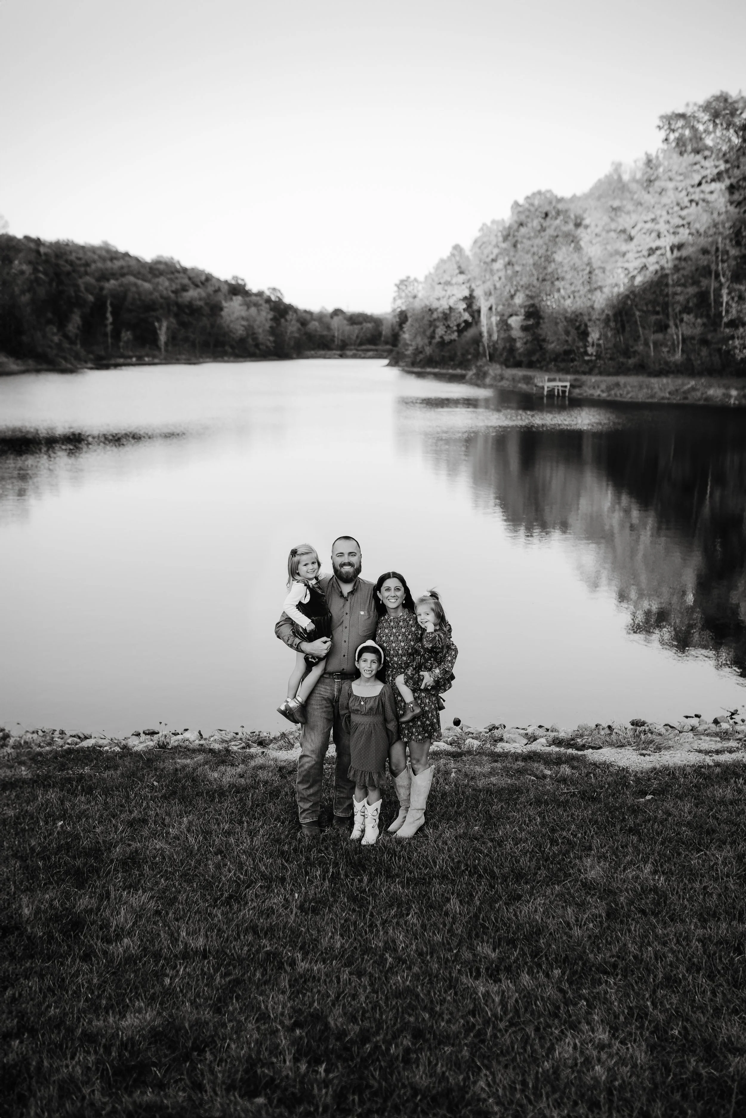 A family of five standing on grass near a lake with trees and hills in the background, all smiling at the camera in black and white.