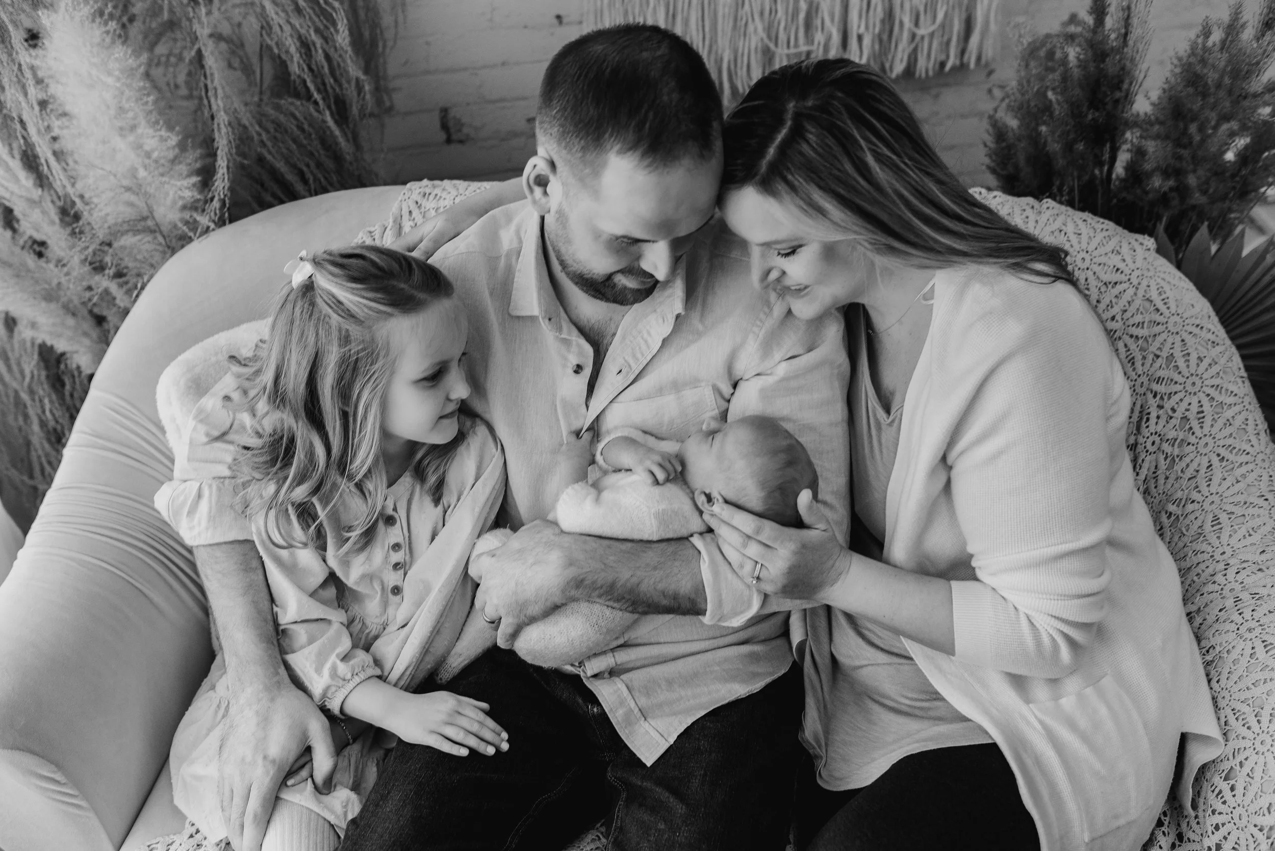 A family of four is gathered on a sofa, with the father holding a newborn baby, the mother leaning in to kiss the baby, and a young girl looking on with a smile.