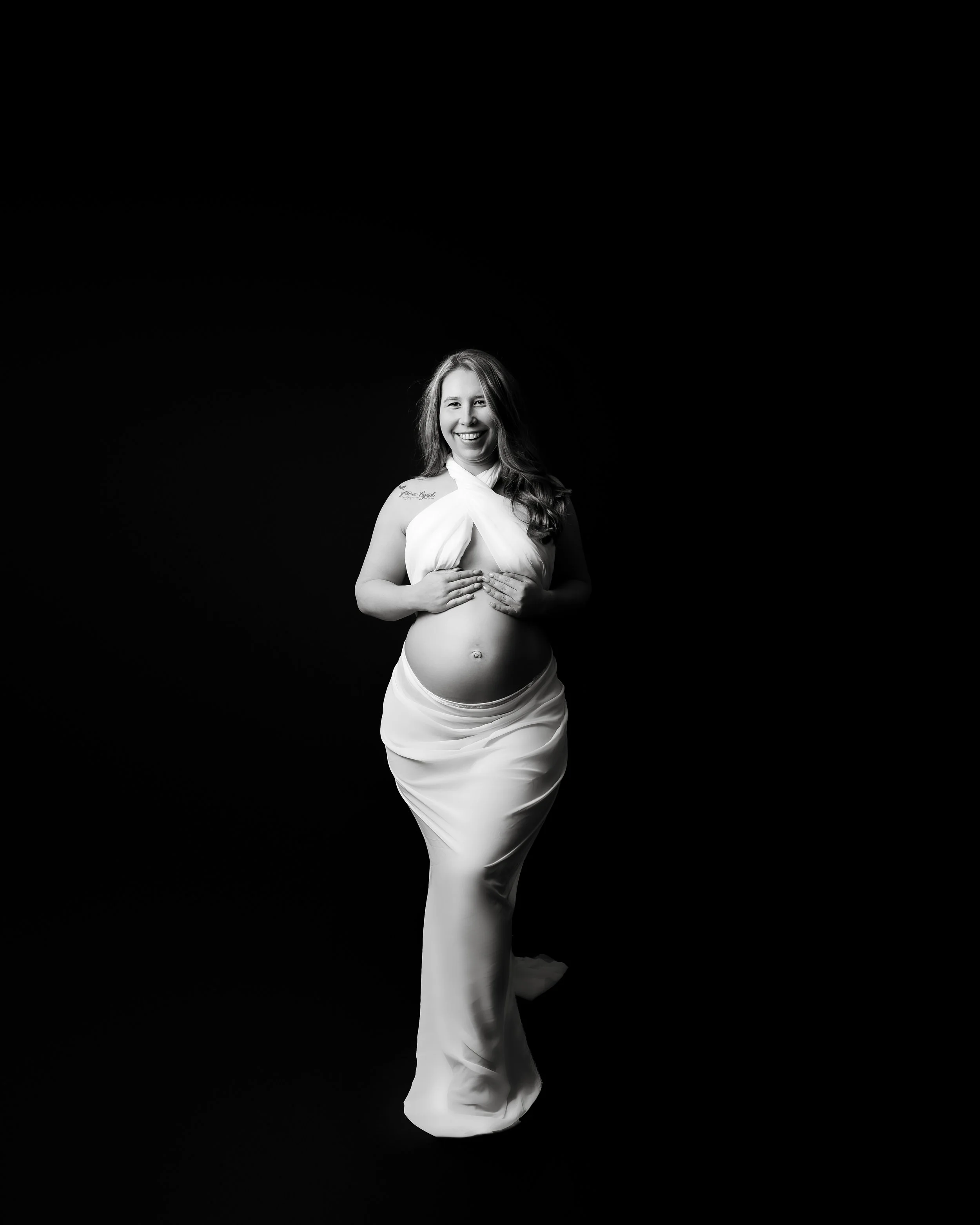 A pregnant woman with long hair, smiling, wearing a white outfit, standing against a black background.