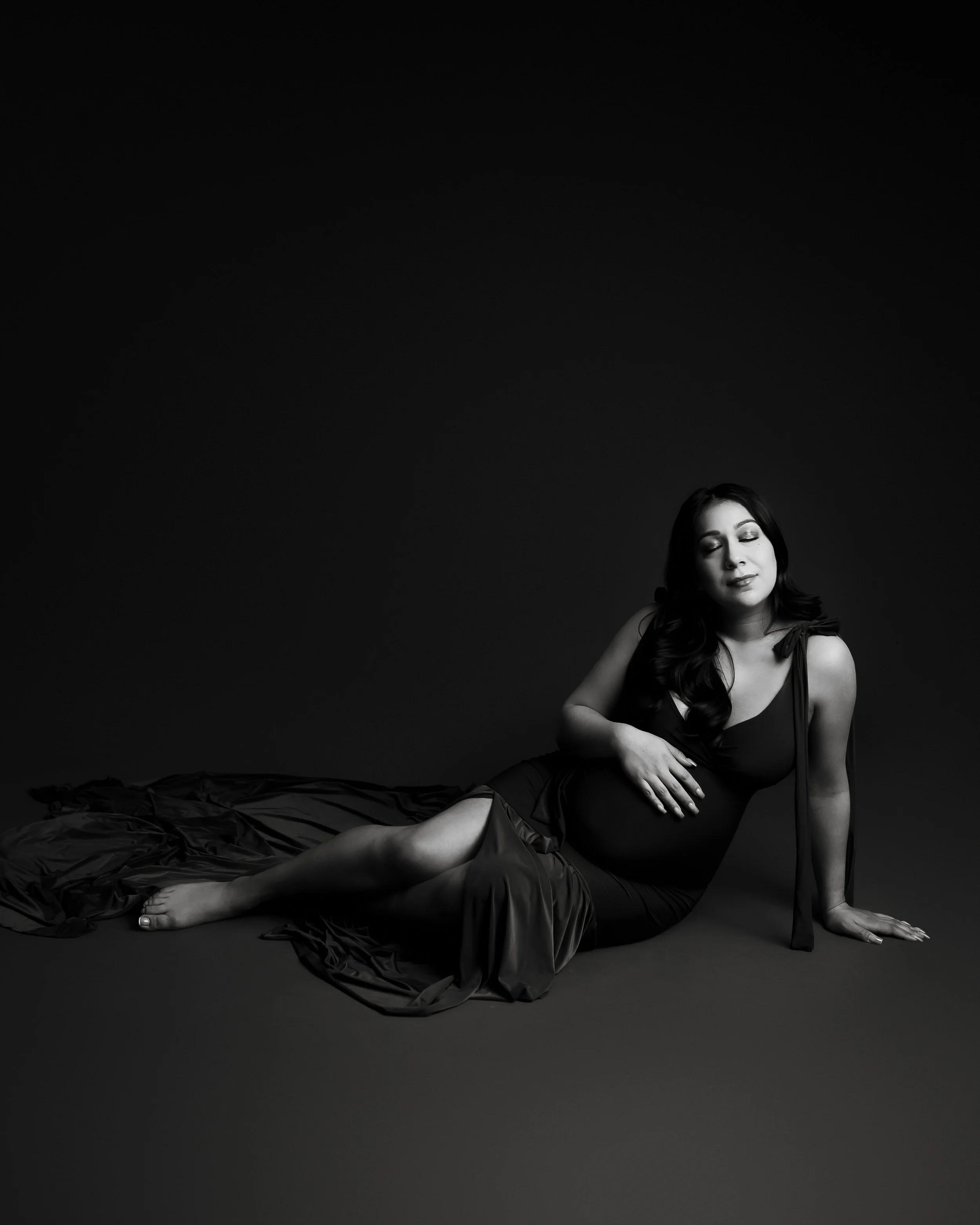 Black and white photo of a pregnant woman sitting on the floor with her eyes closed, wearing a dark dress and a long fabric draped behind her.