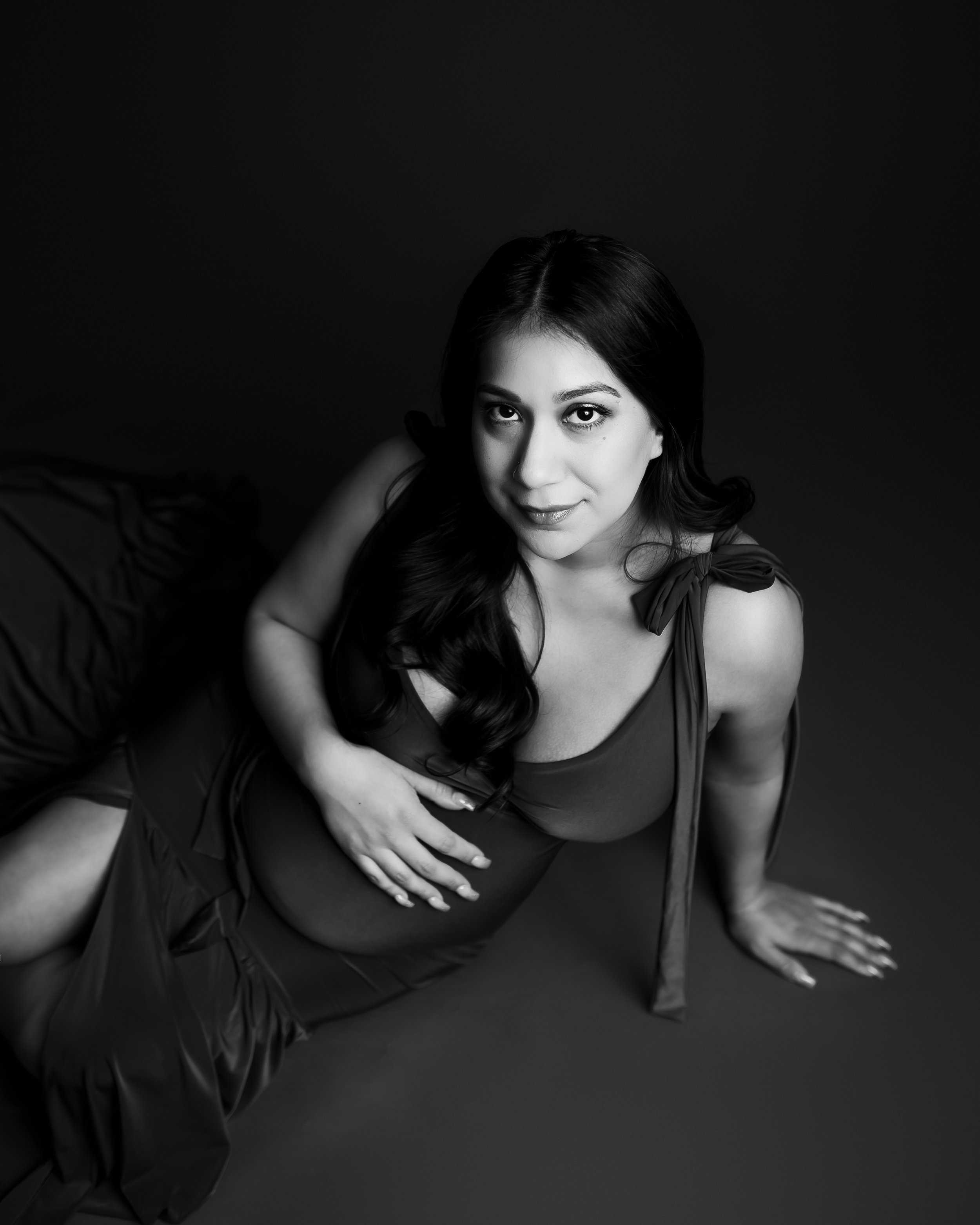 A young woman with long dark hair and light skin posing on the floor in a black dress with shoulder ties, looking at the camera with a slight smile, in a black and white studio portrait.