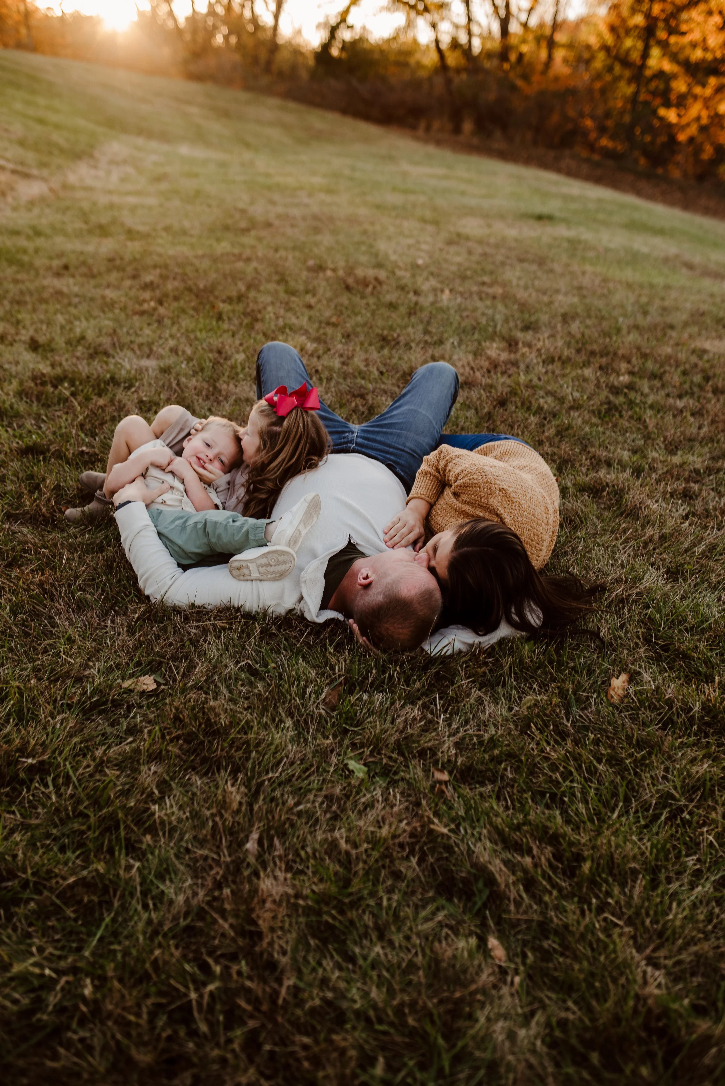Family of four lying on the grass and cuddling in a park during sunset fall, with orange trees in the background.