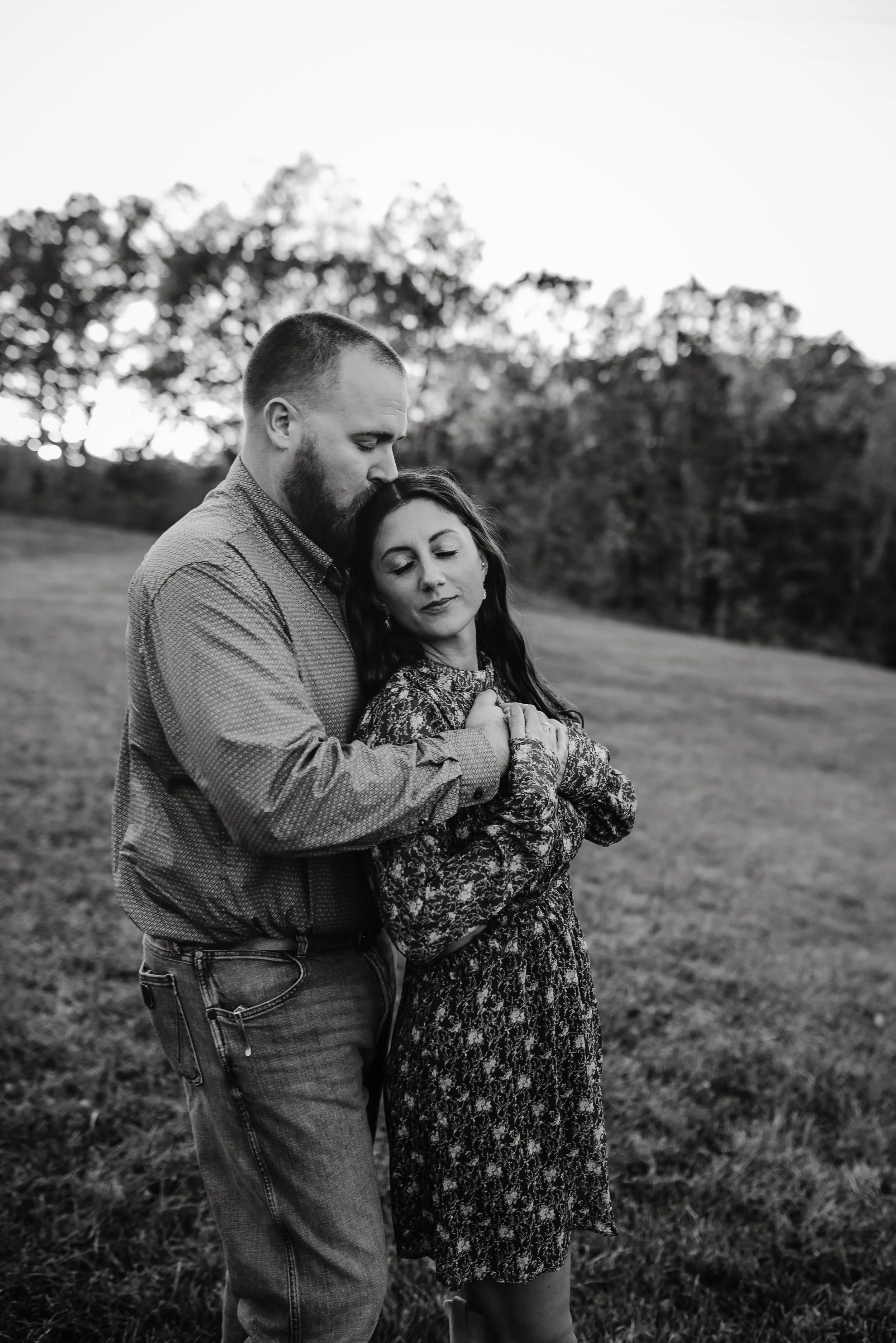 A black-and-white photo of a couple standing outdoors in a field, embracing each other with eyes closed, with trees in the background.