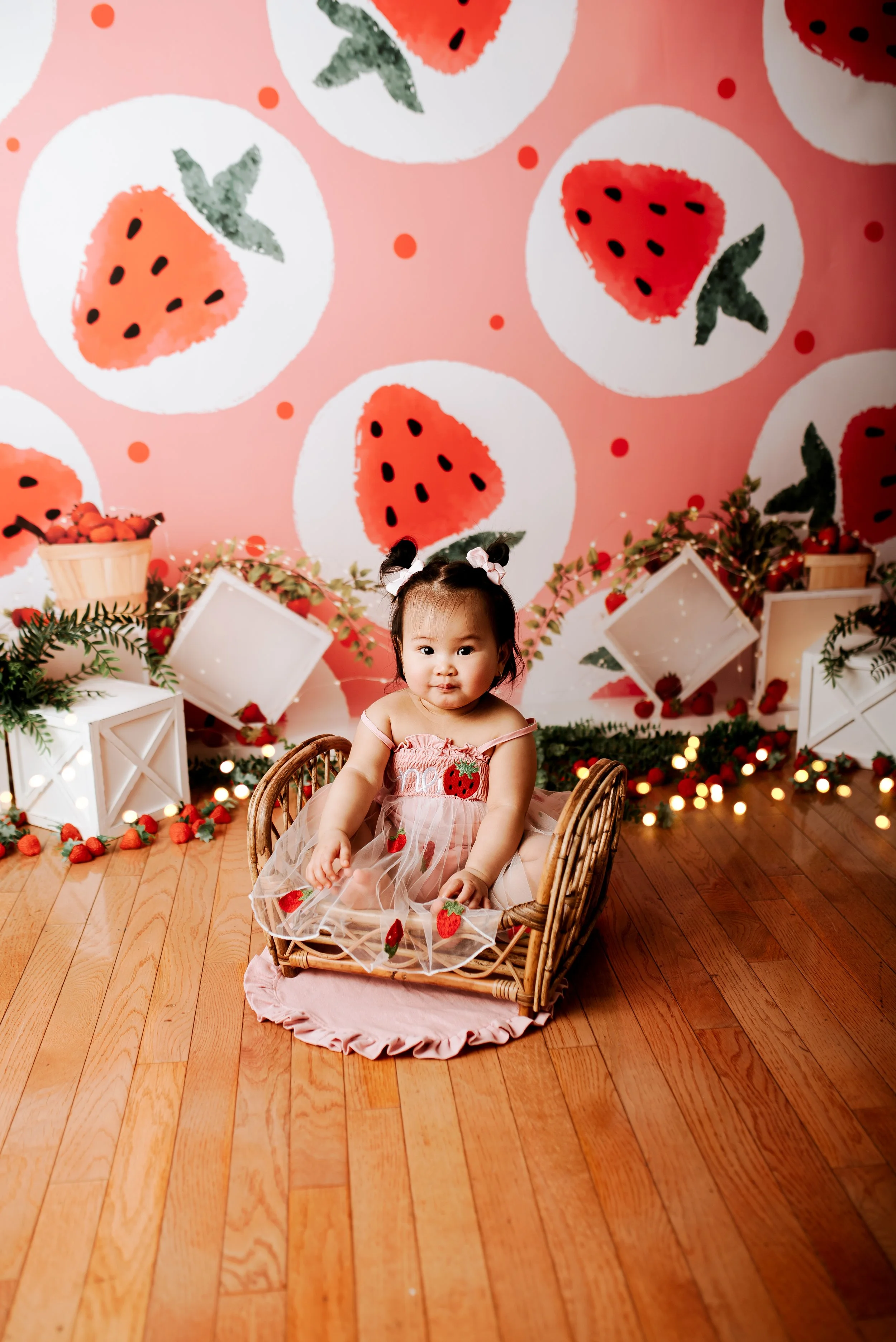 A young girl dressed in a pink dress with strawberry patterns, sitting in a wicker basket, in front of a strawberry-themed backdrop with strawberries, greenery, and string lights.
