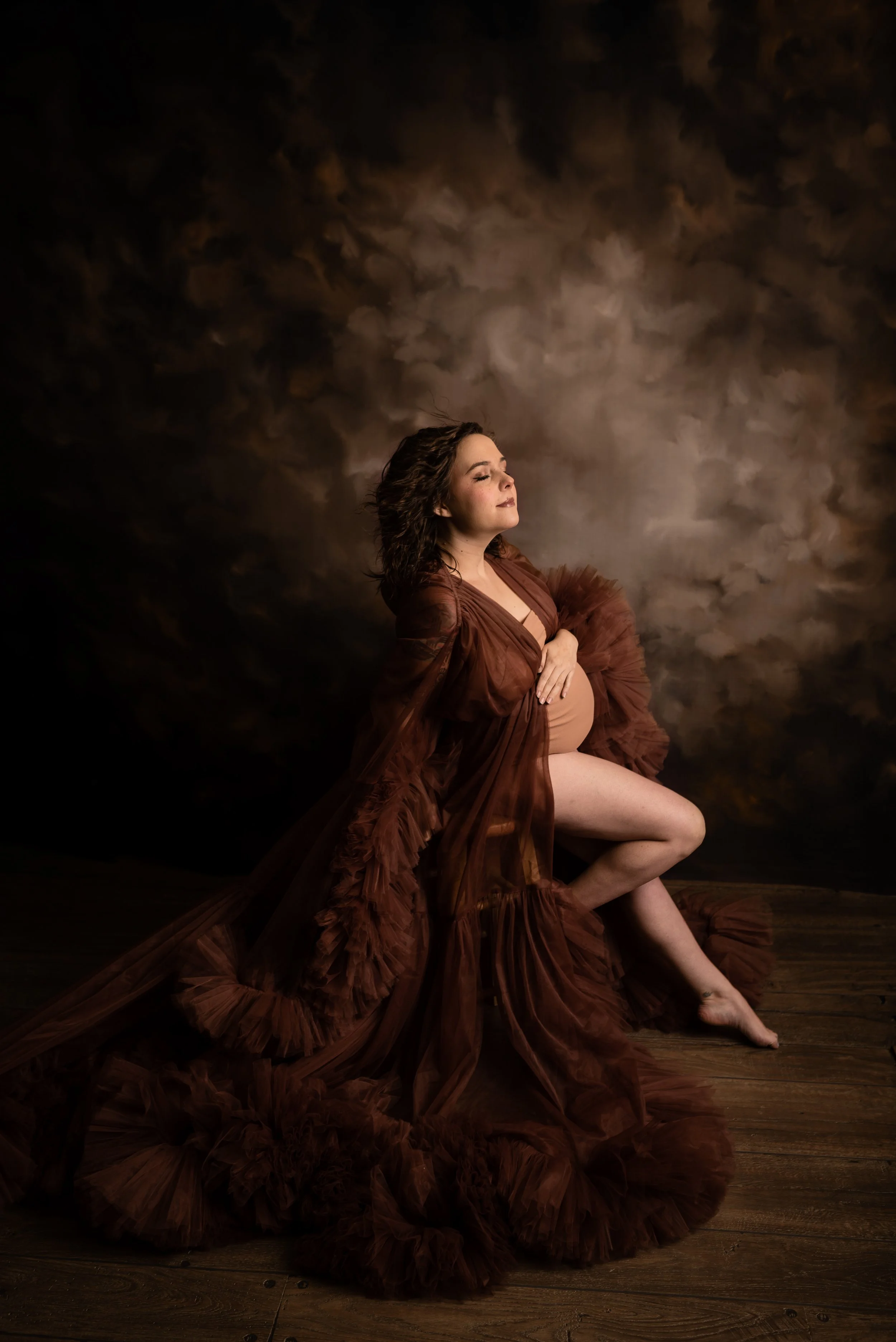A pregnant woman in a brown gown sitting on a wooden floor against a dark, cloudy background, with her eyes closed and hands resting on her belly.