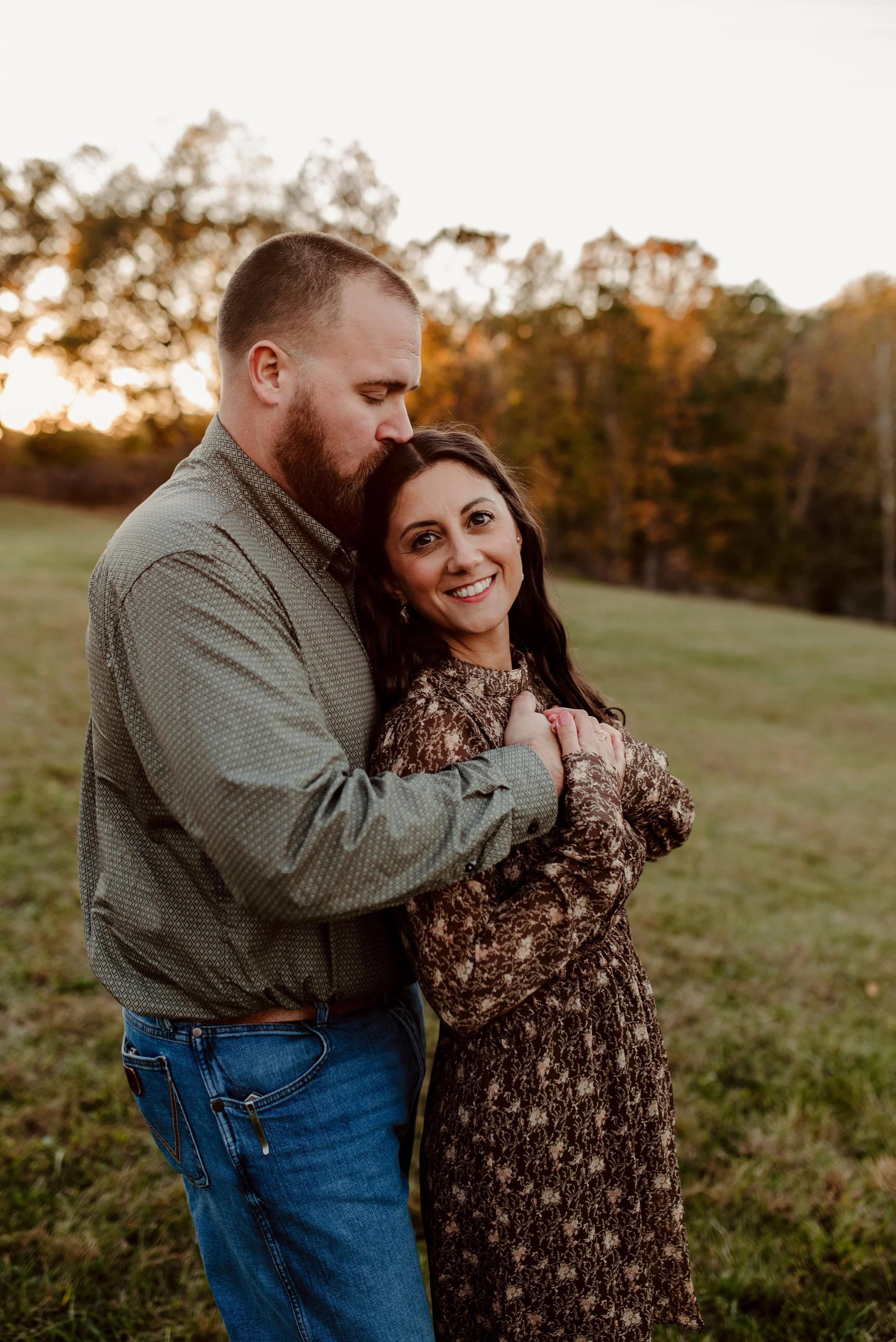A couple standing together outdoors at sunset, with the man kissing the woman's head and the woman smiling at the camera, in a grassy area with trees in the background.