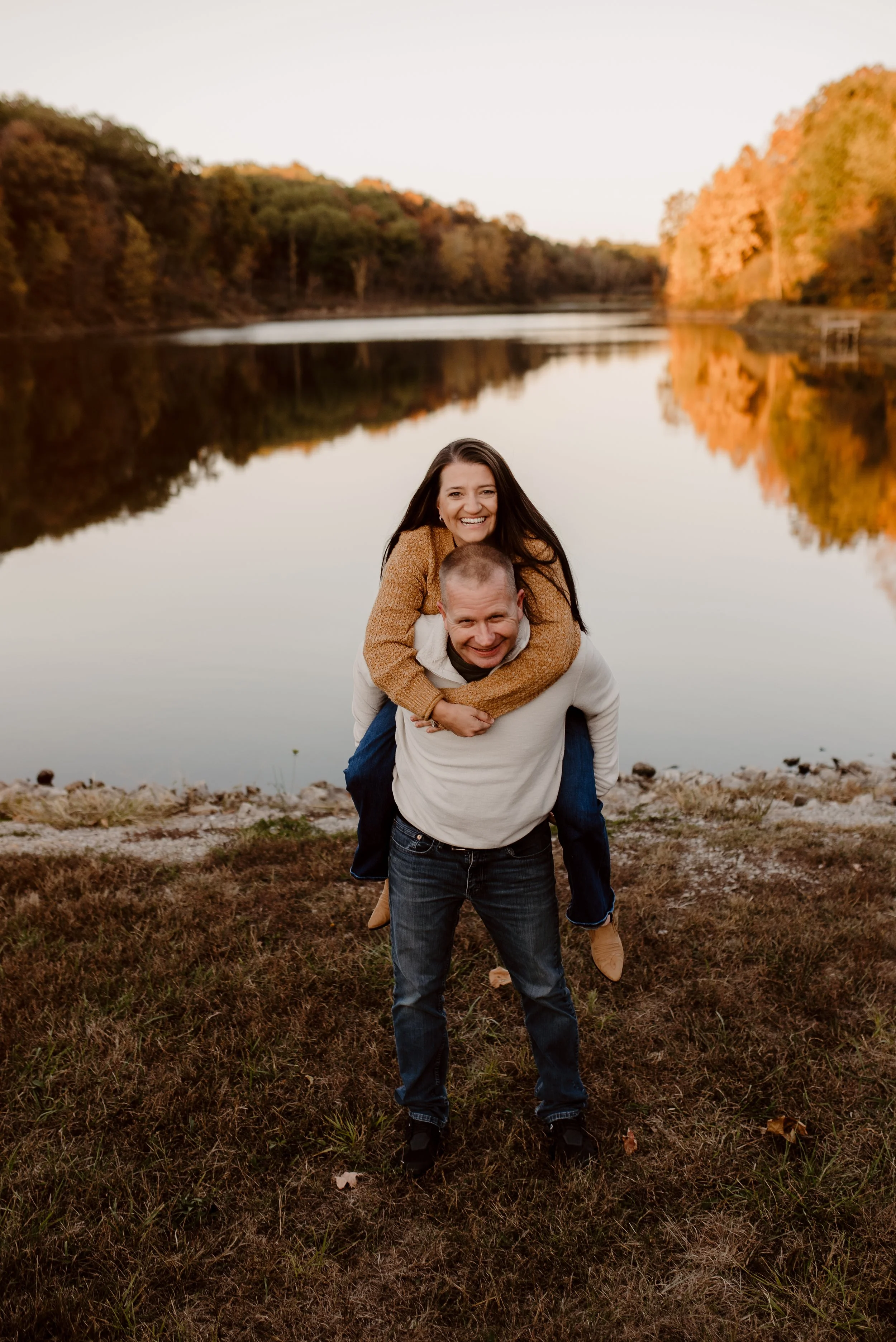 A couple standing outdoors near a lake during autumn, with the woman on the man's back, both smiling happily, surrounded by colorful fall foliage.