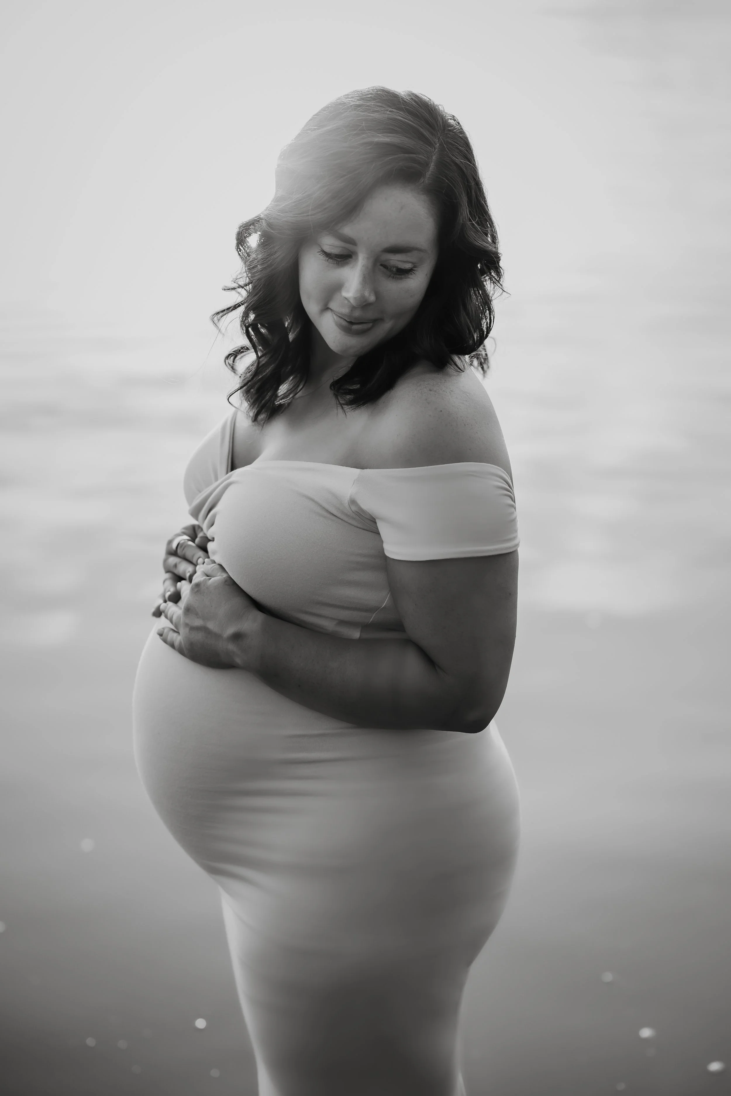 Black and white photo of a pregnant woman standing by the water, gently holding her belly with both hands, with soft light illuminating her face and hair.