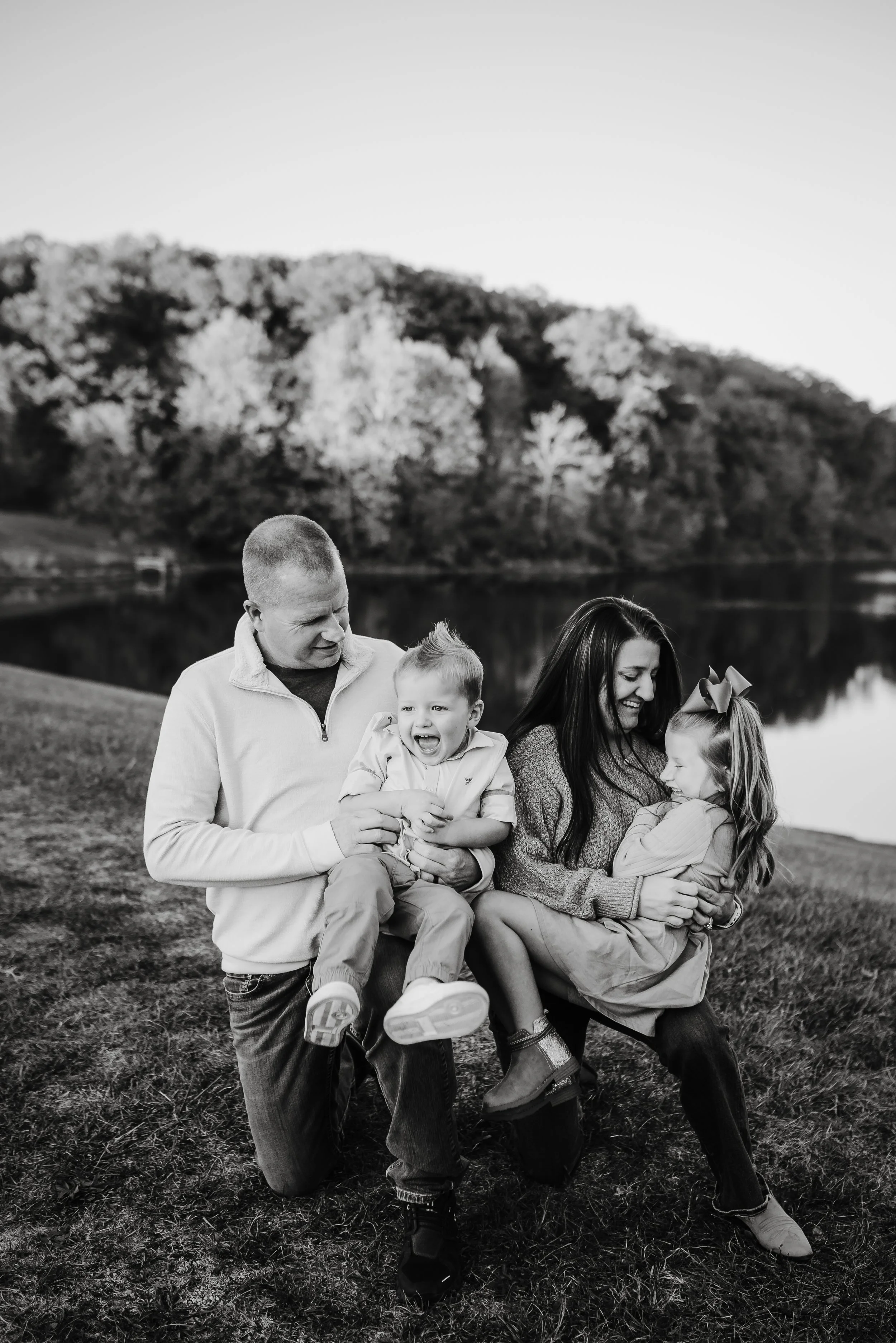 A black and white photo of a family of four outdoors beside a lake. The father, kneeling, holds a laughing young boy, while the mother, also kneeling, holds an amused young girl with a bow in her hair. Trees and hills are in the background.