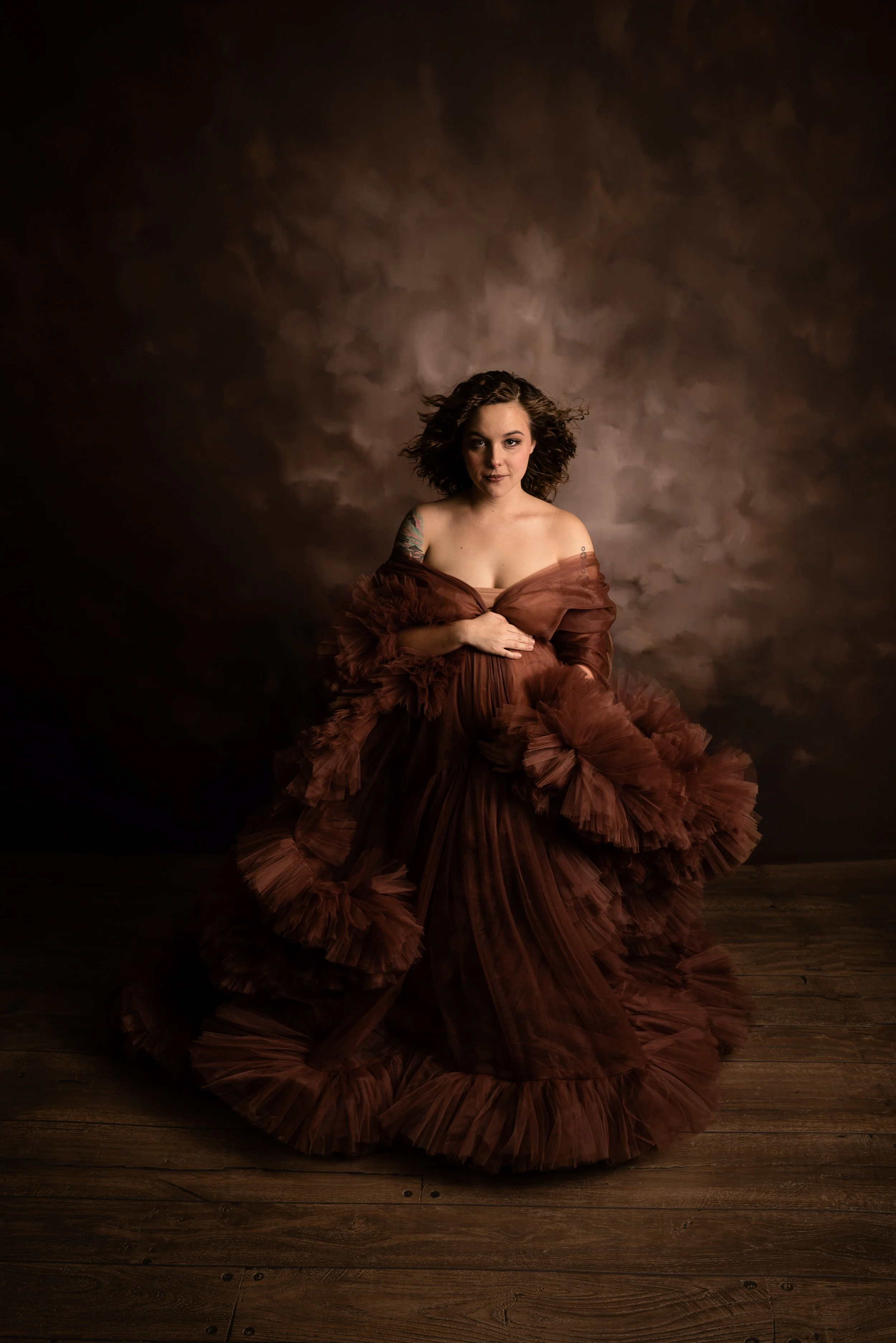 A woman with curly hair wearing a voluminous, ruffled brown gown stands on a wooden floor against a dark, cloudy background.