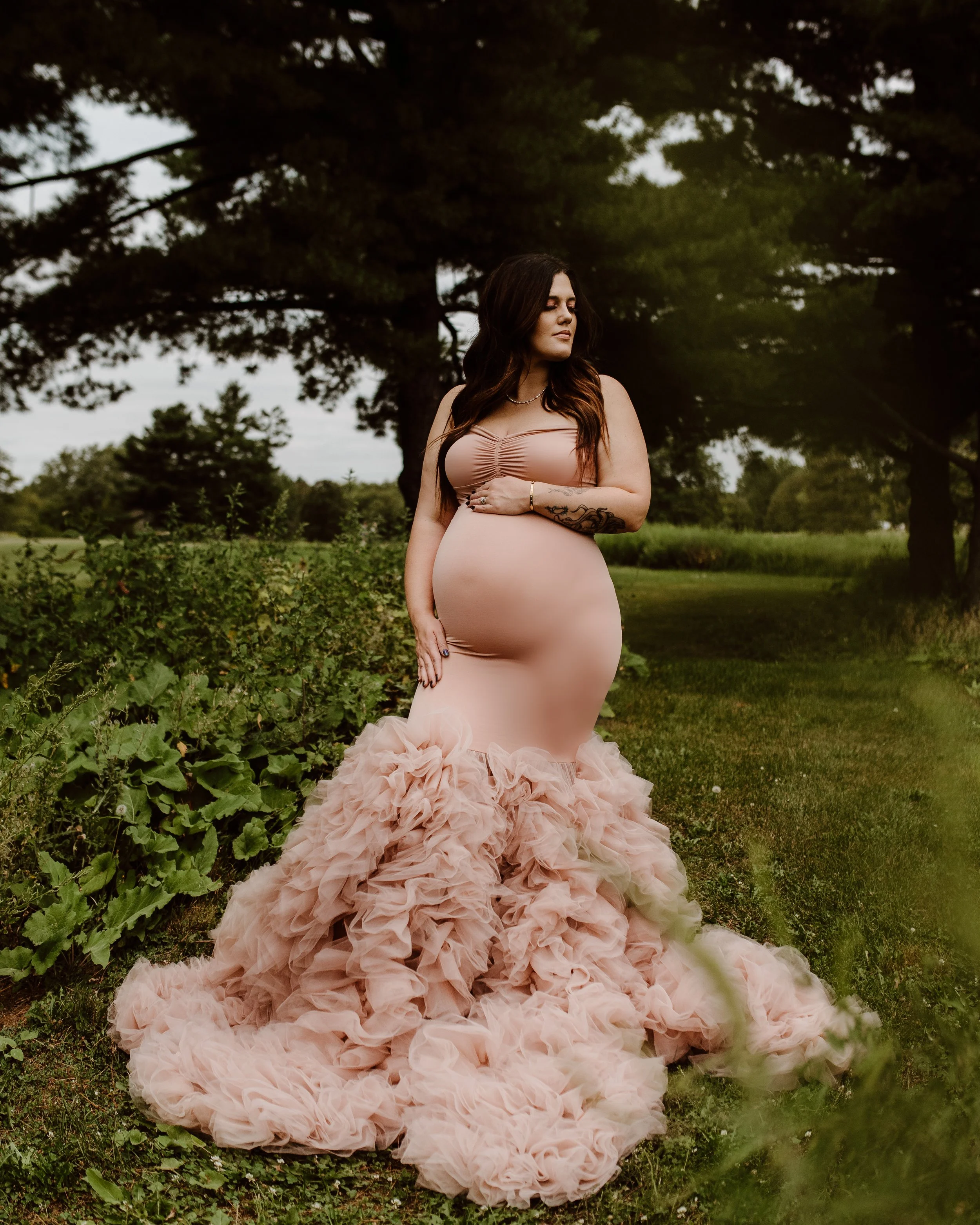 A woman wearing a nude-colored, strapless gown with a ruffled, tulle train standing in a grassy outdoor setting with trees in the background.