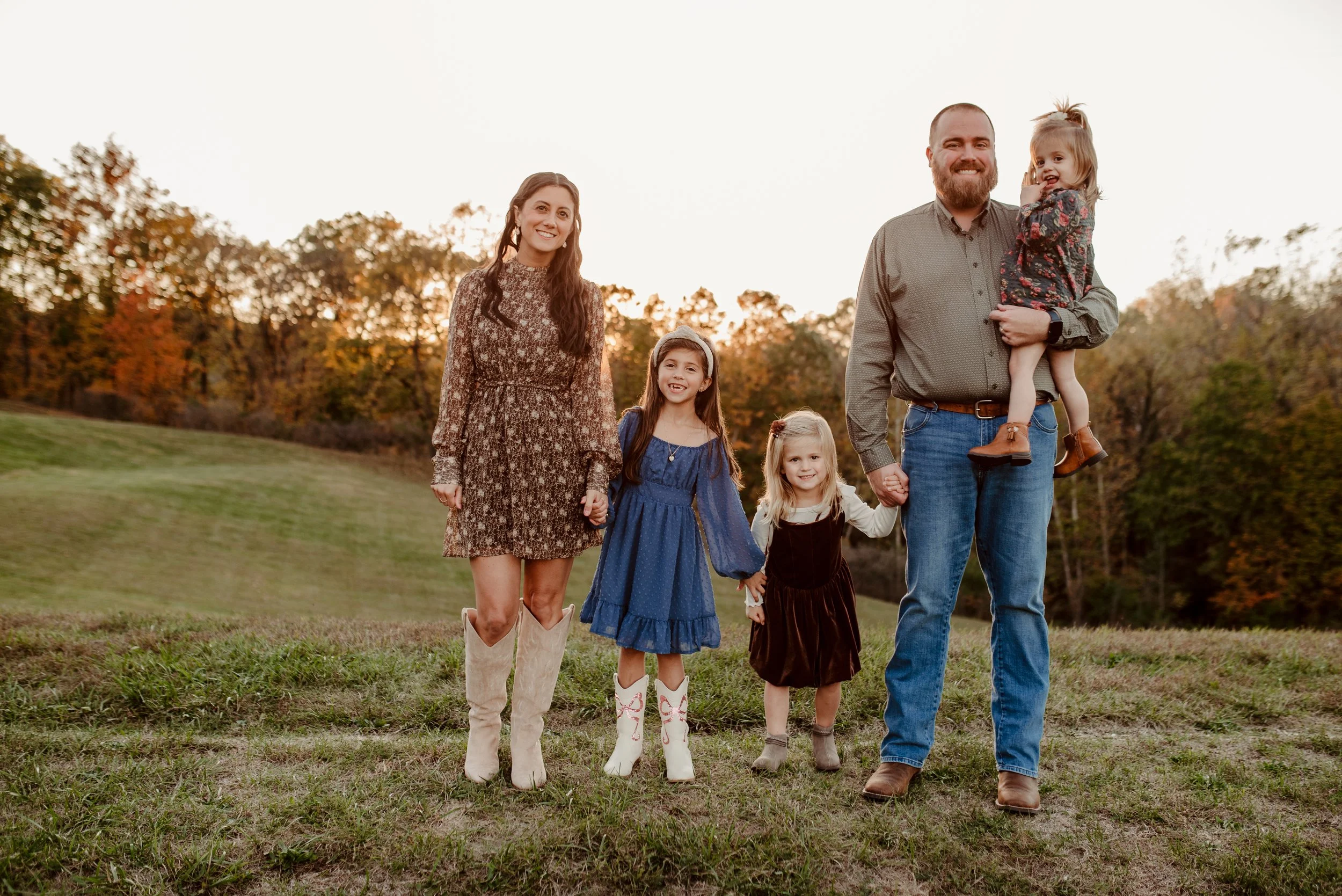 Family of five walking outdoors during sunset, holding hands, with trees and hills in the background, dressed in fall clothing.