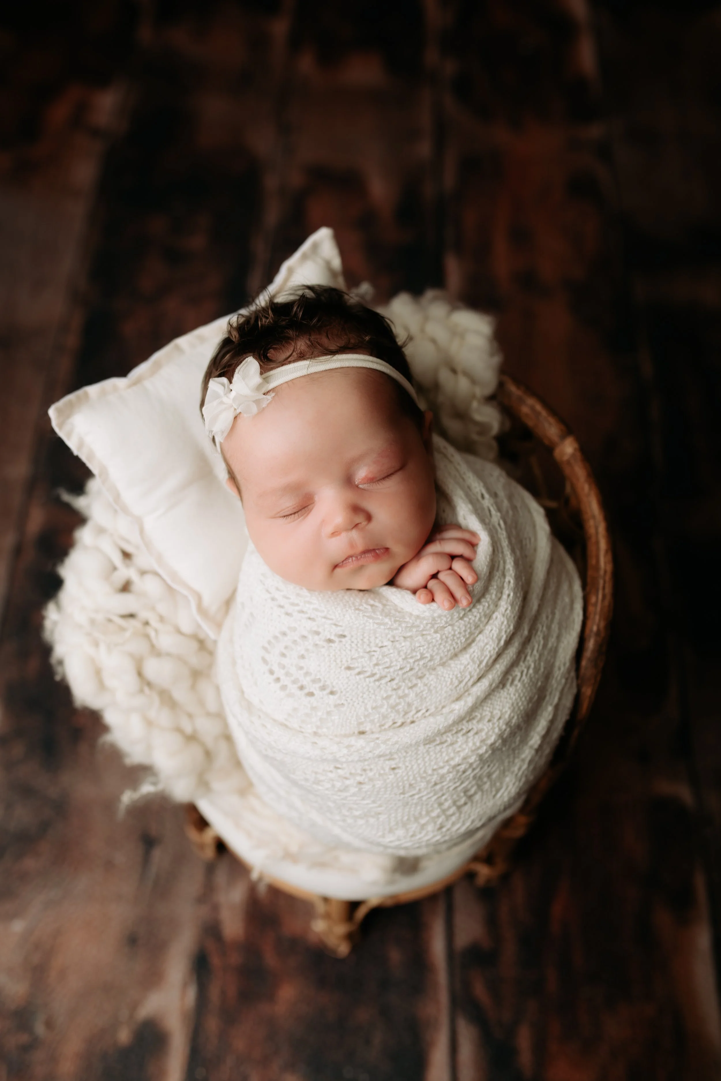 A newborn baby girl asleep, wrapped in a white knitted blanket, with a matching headband, lying on a small pillow in a wicker basket on a dark wooden floor.