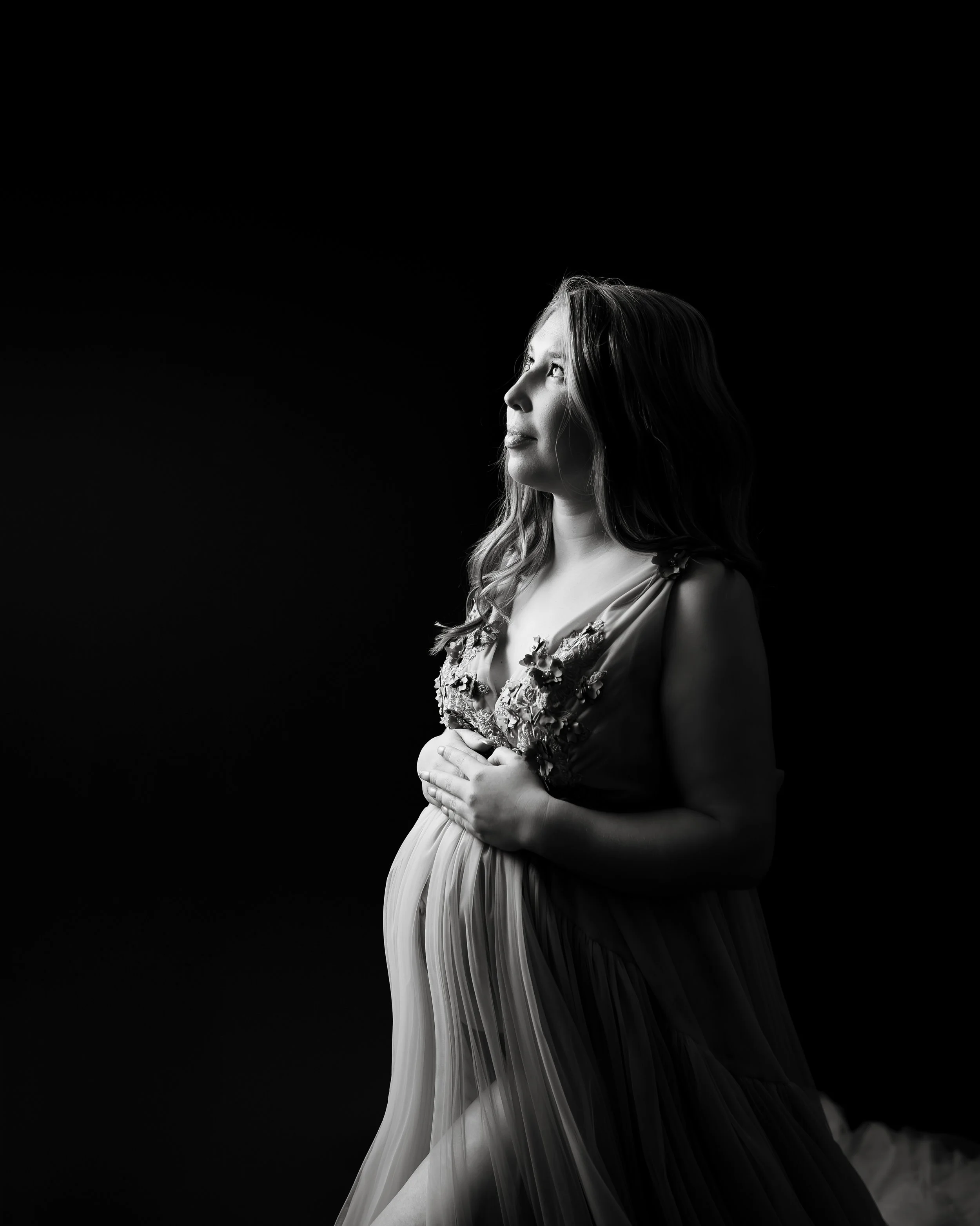 A black and white photo of a pregnant woman in a flowing dress, holding her belly, looking upward, against a dark background.