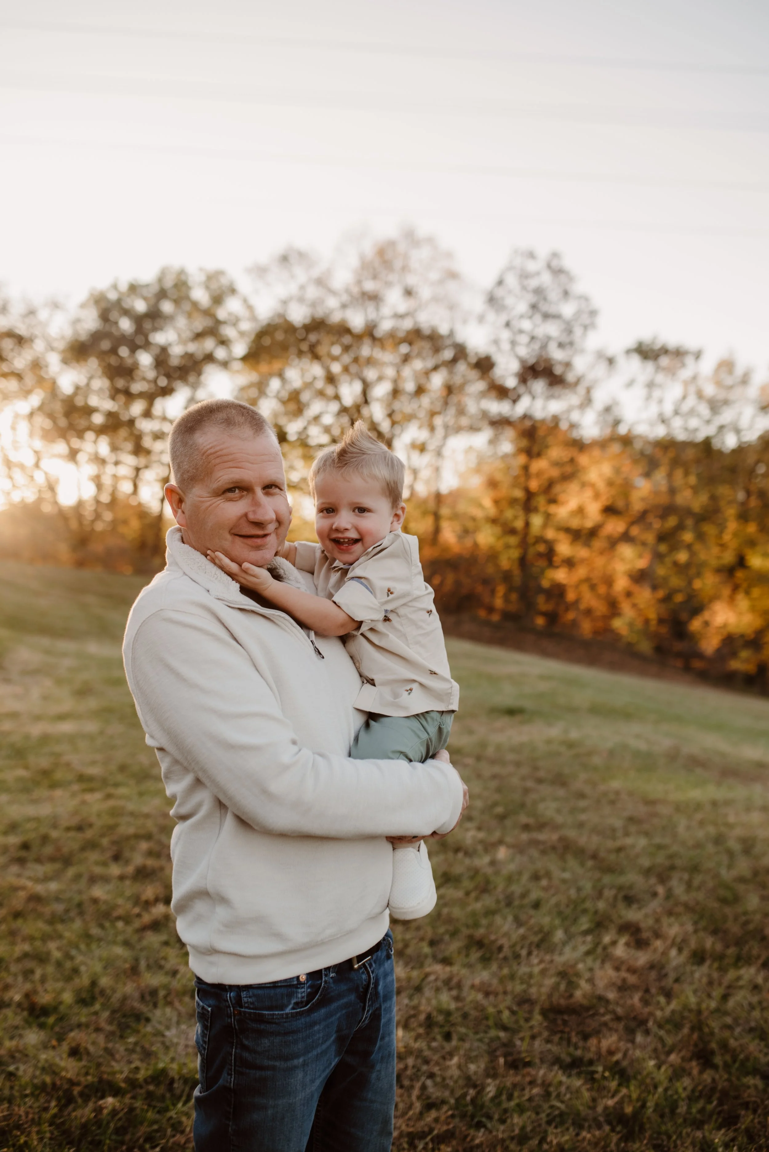 Father holding his young son outdoors during autumn sunset with colorful fall foliage in the background.