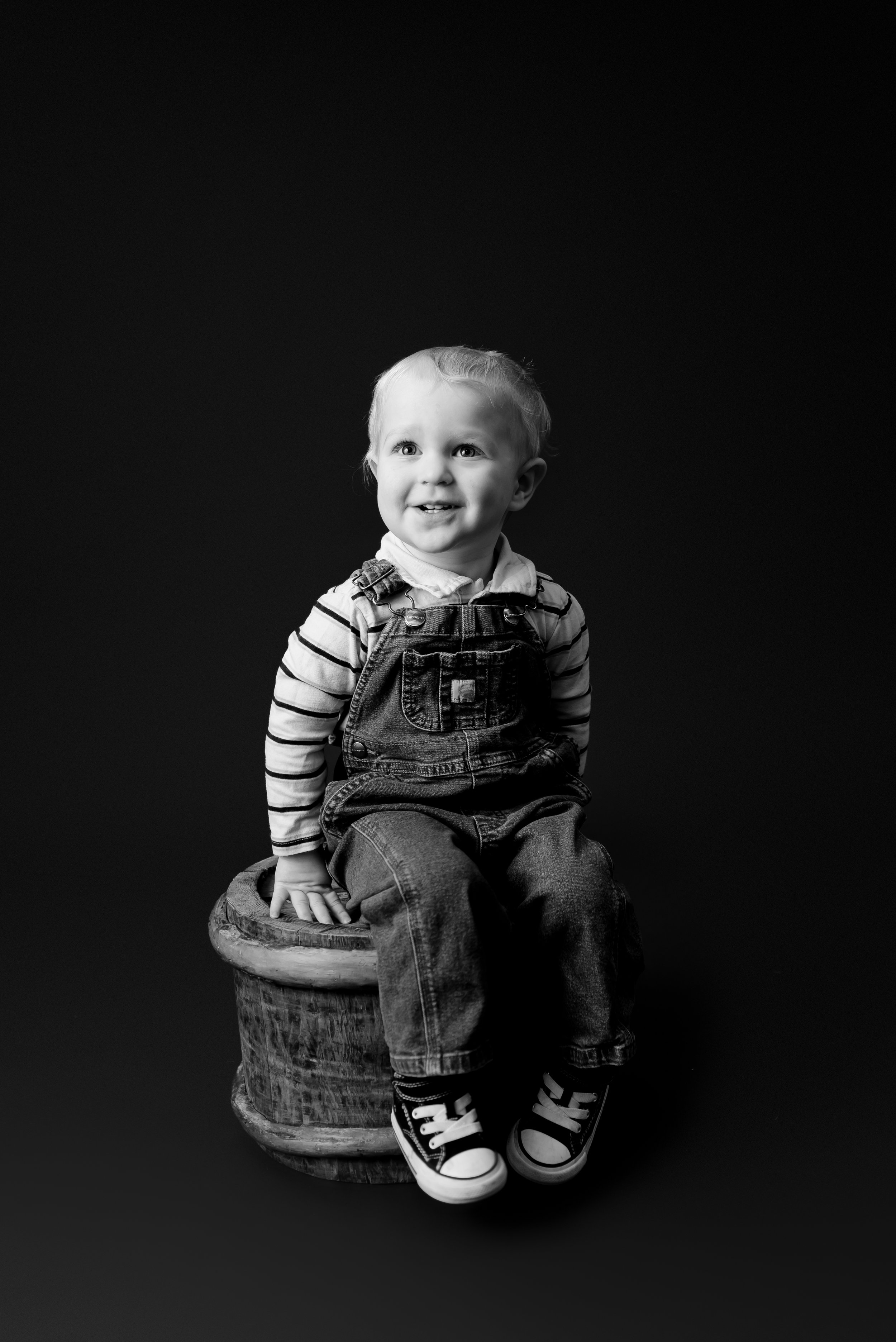 A young child sitting on a wooden stool, smiling, wearing striped long-sleeve shirt, denim overalls, and sneakers, in a black and white studio portrait.