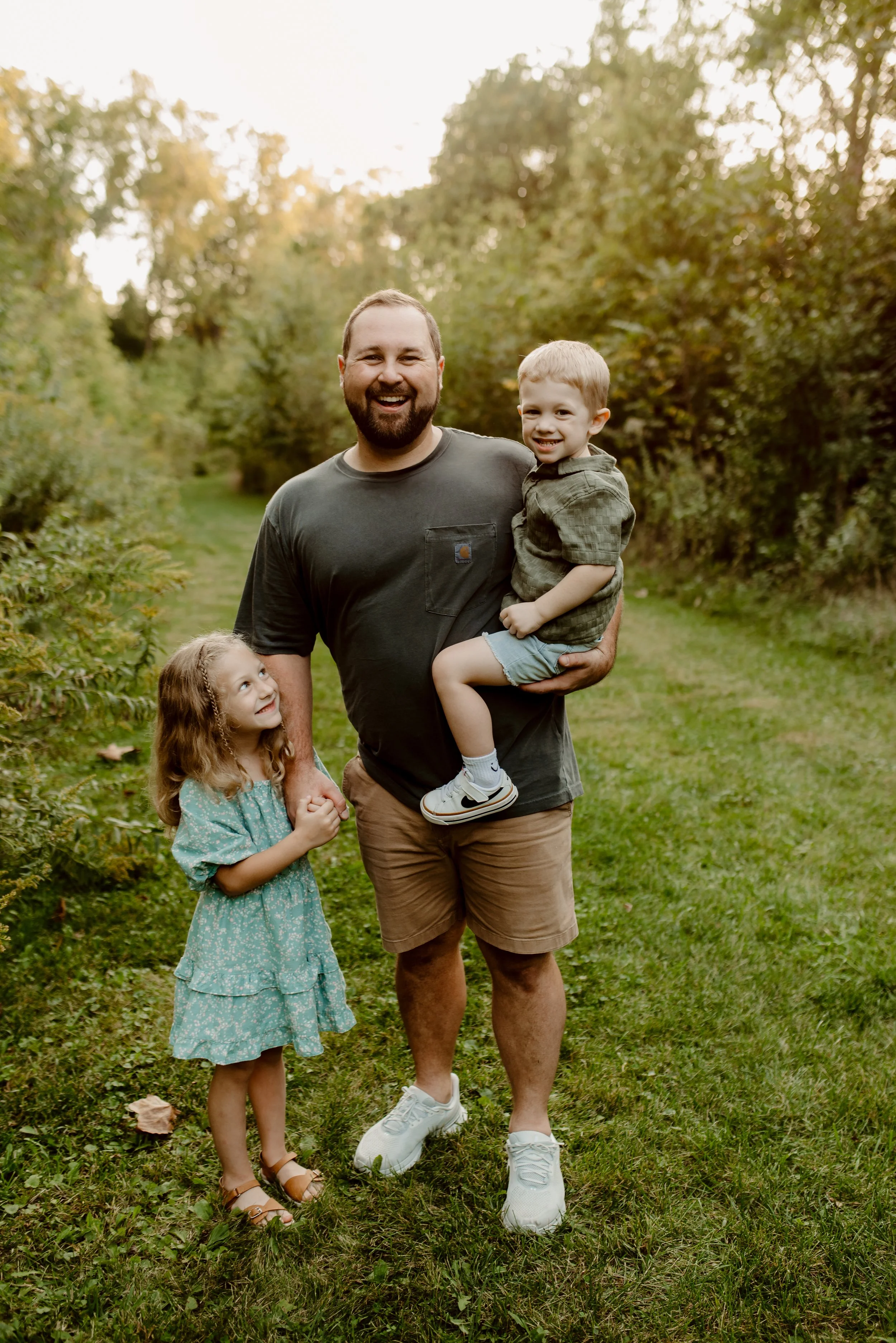 A man holding a young boy in his arms, standing on a grassy path surrounded by trees, with a young girl looking up at him, all smiling in a park during daytime.