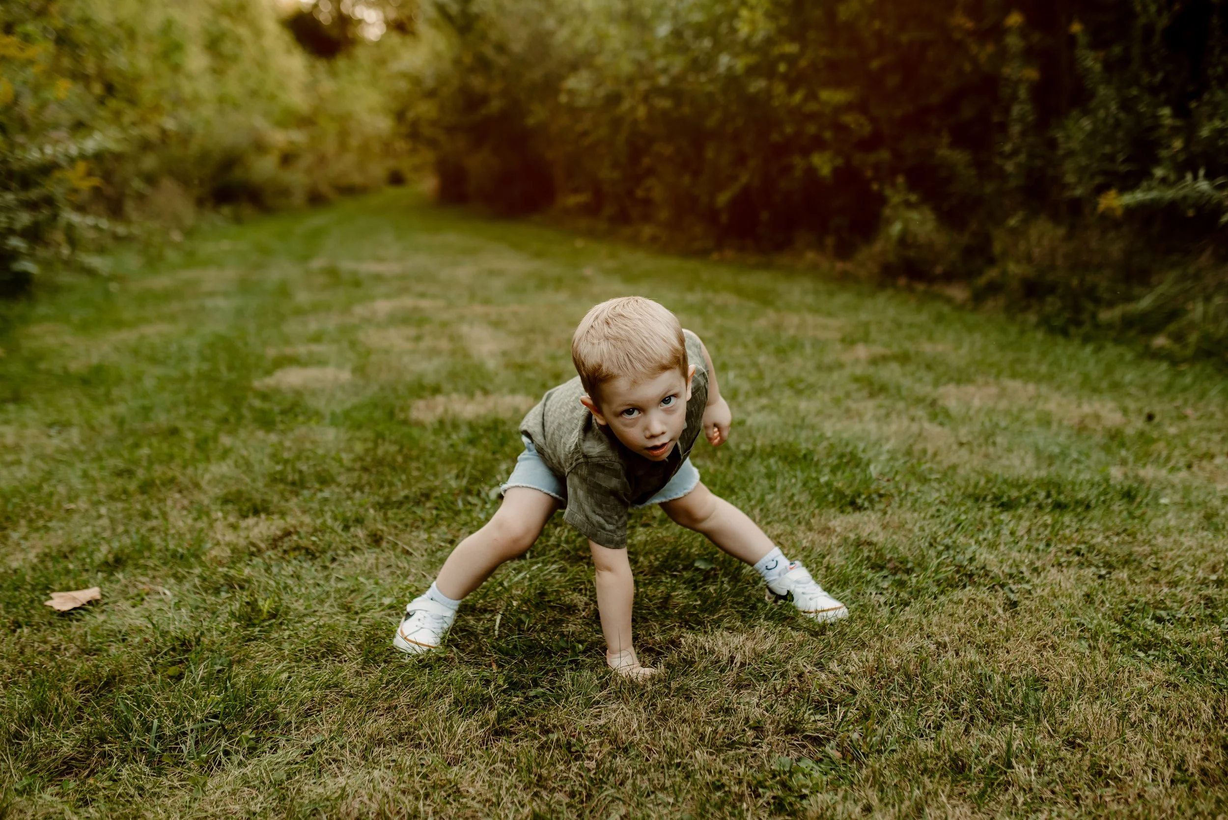 A young boy in a gray t-shirt and shorts crouching on a grassy path surrounded by trees, looking directly at the camera with a serious expression.