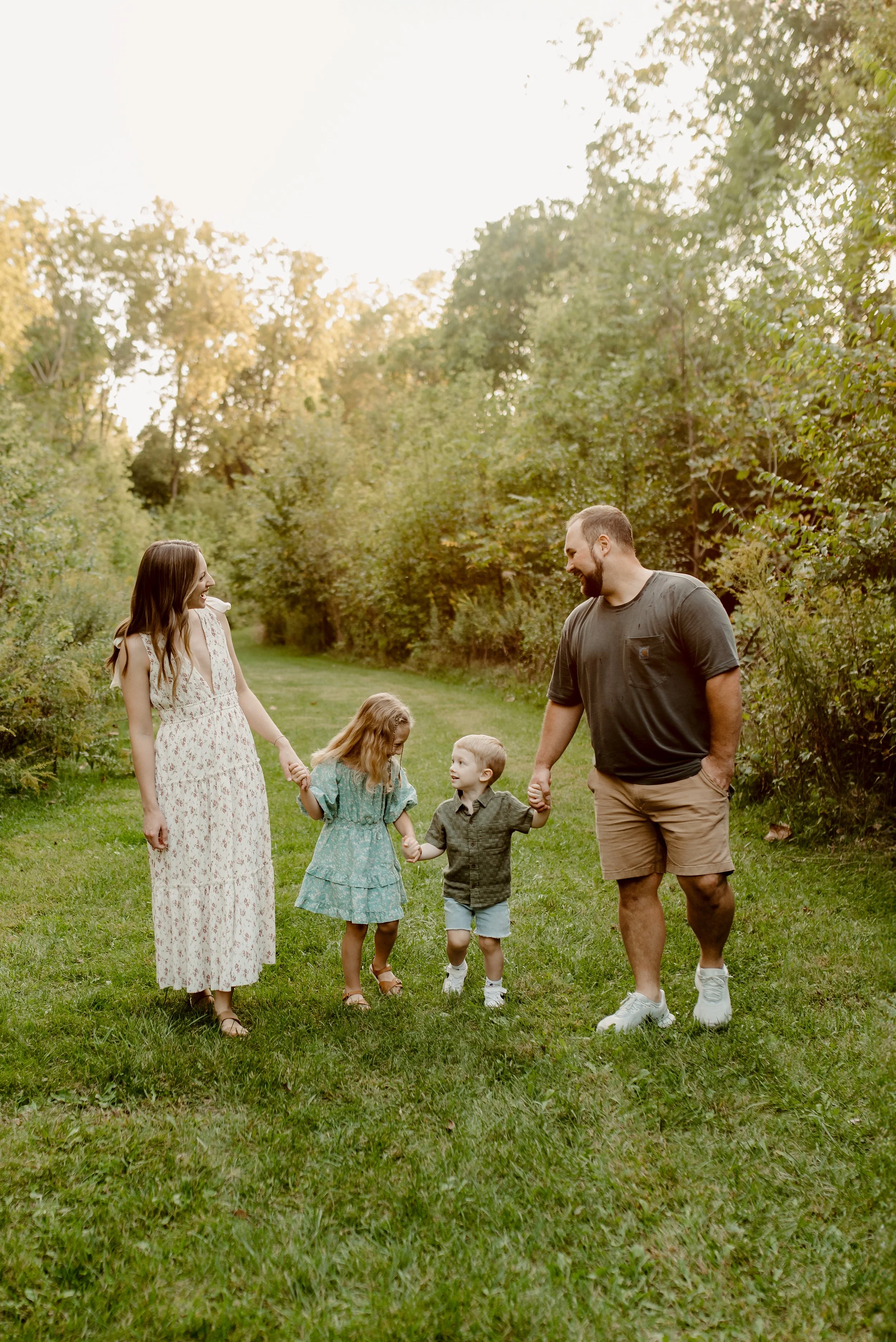 A family of five walking hand-in-hand outdoors on a grassy trail through a wooded area, enjoying each other's company and smiling at each other.