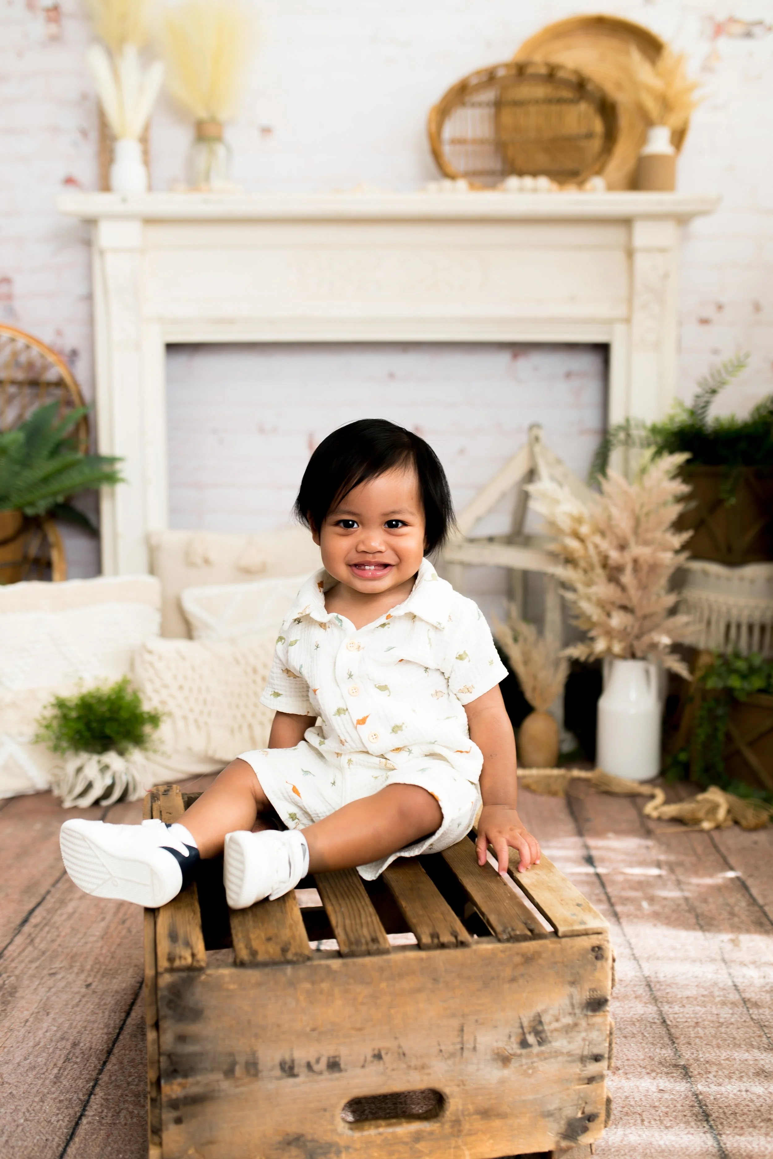 A young child sitting on a wooden crate in a cozy, decorated room with plants and a white fireplace.