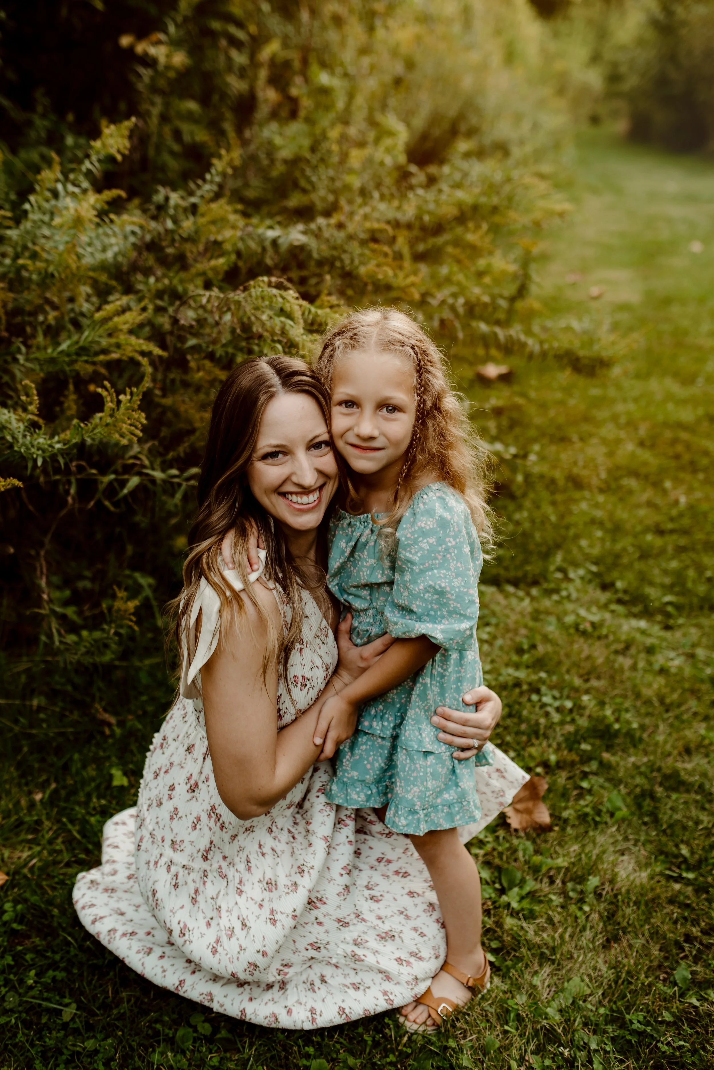 A woman and a young girl hugging outdoors on a grassy path with greenery in the background.