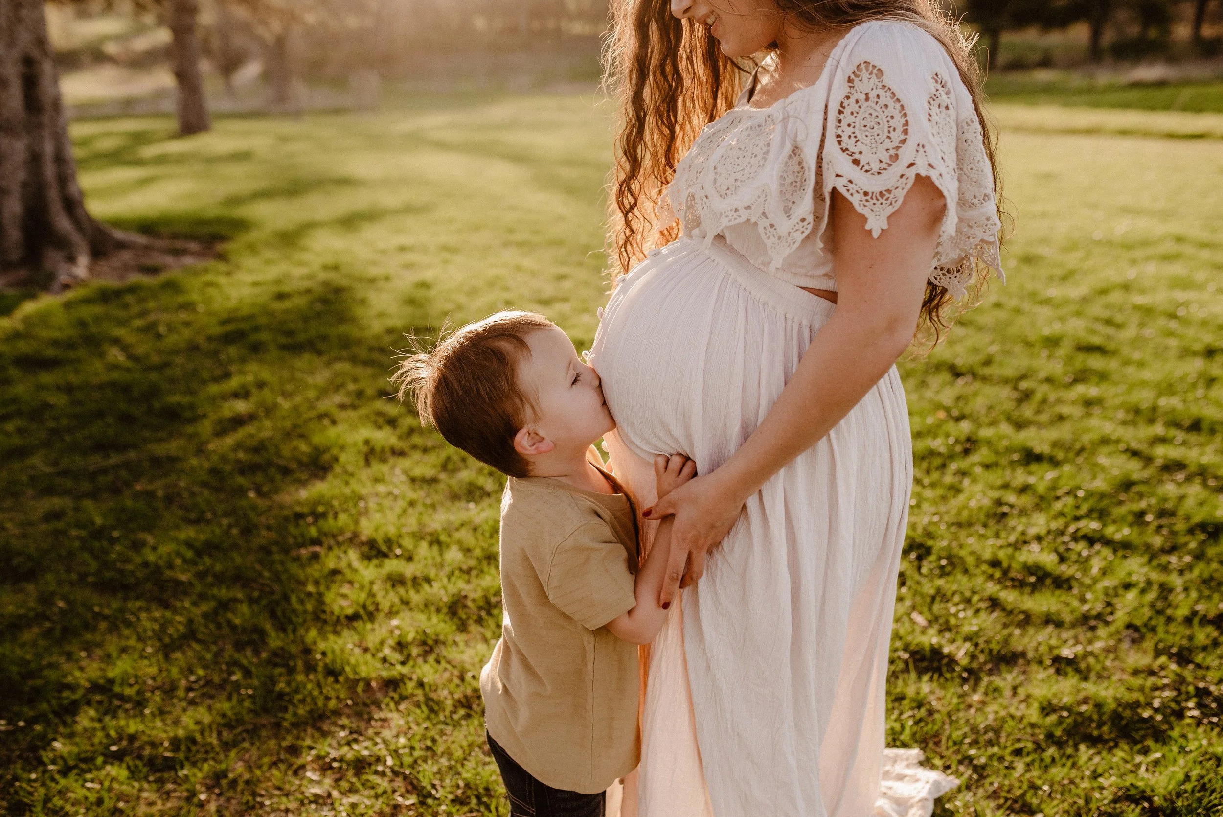 A young boy kisses a pregnant woman on her belly during a sunny outdoor scene in a park.