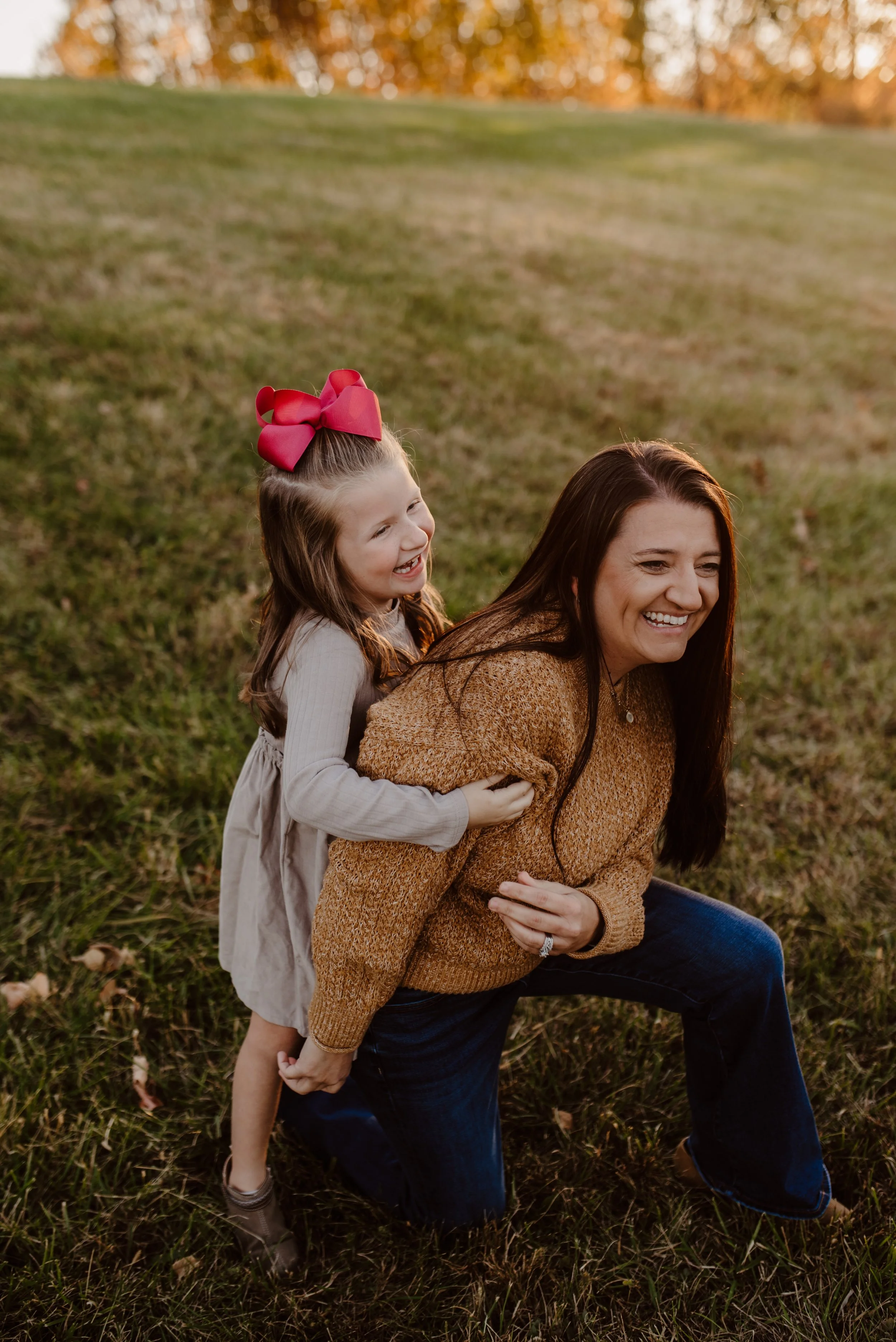 A woman and a young girl playing outside on a grassy field during sunset. The girl with a big pink bow on her head is riding on the woman's back, both smiling and enjoying the moment.