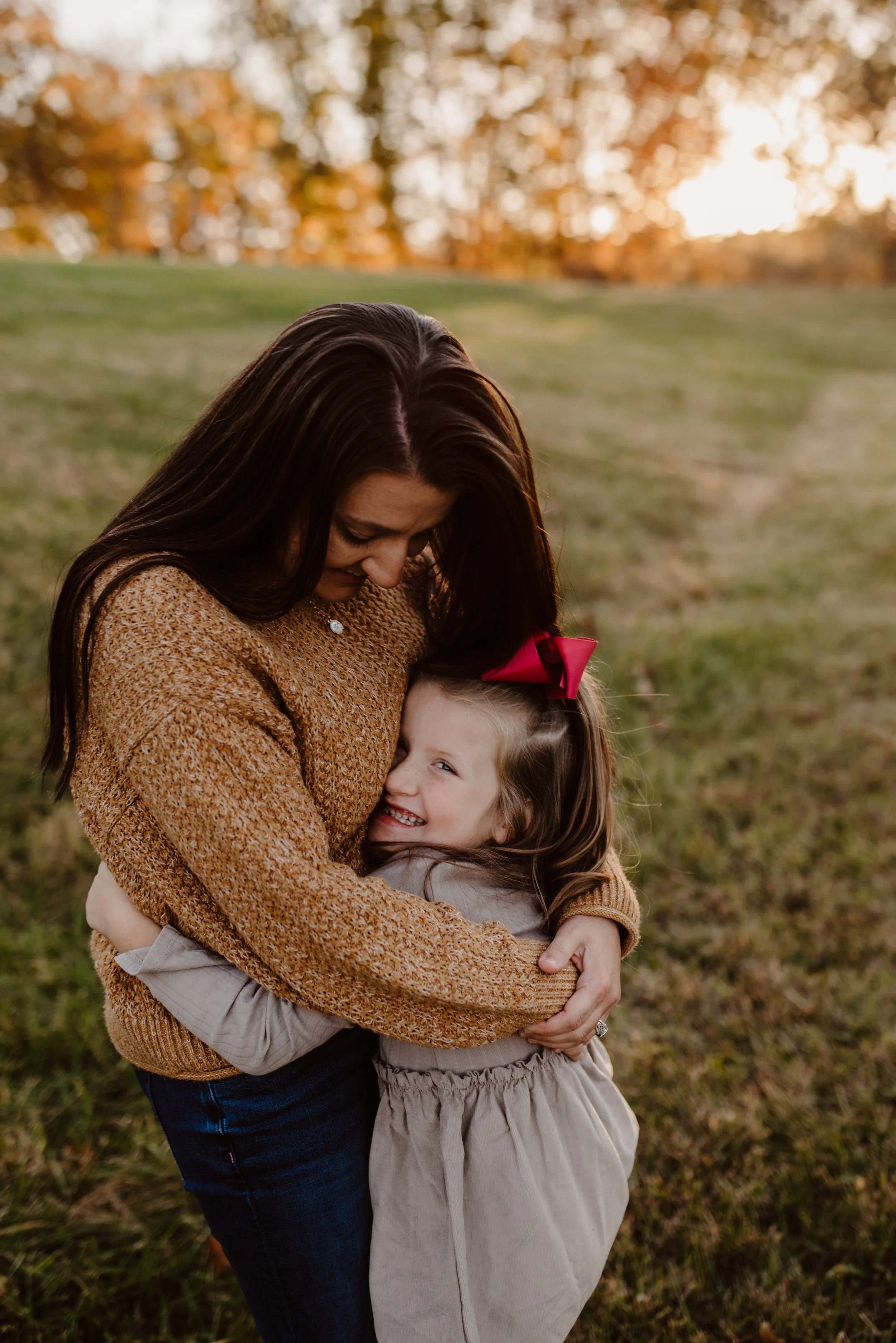 A woman and young girl hugging outdoors during sunset, smiling with trees in the background.