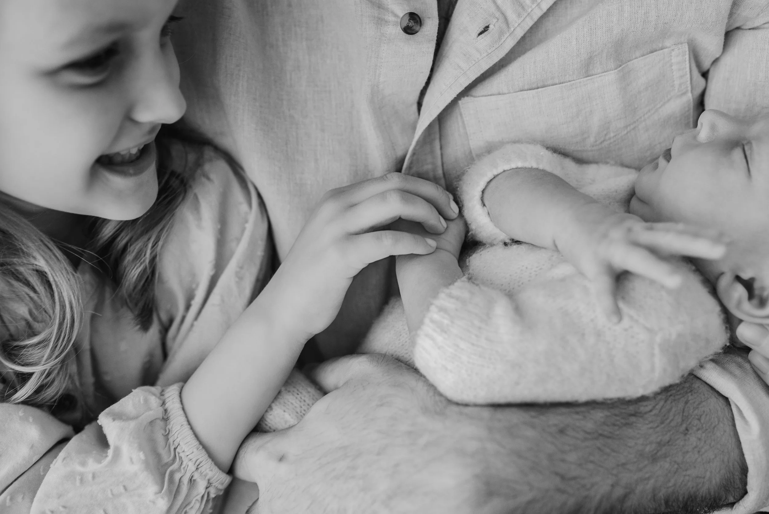 A young girl smiling and touching a baby's hand while the baby is breastfeeding. The girl has long hair and is wearing a patterned top. The baby is lying on a mother's chest, wearing a sweater.