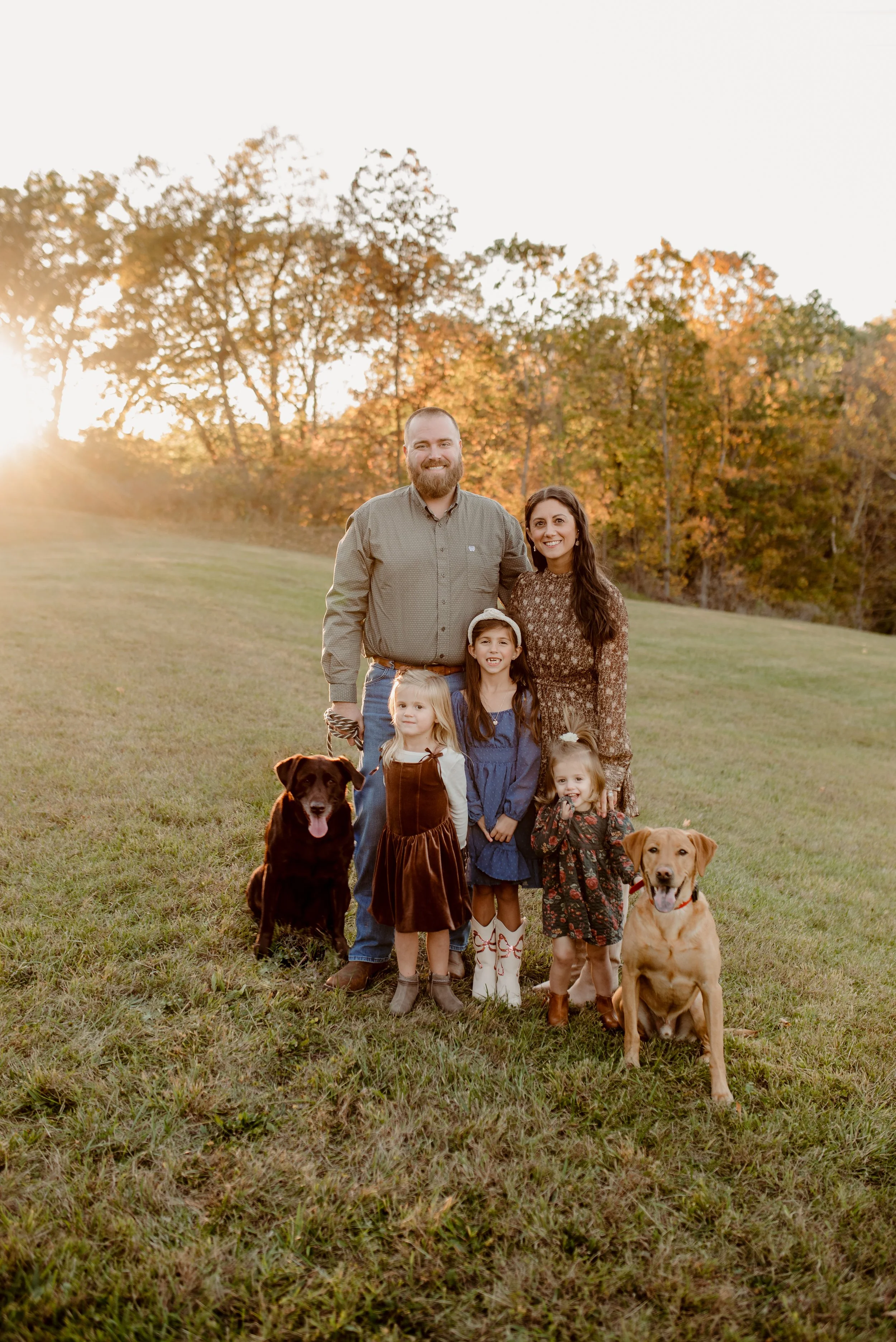 A family of six, including two dogs, standing outdoors on grass during sunset with trees in the background.