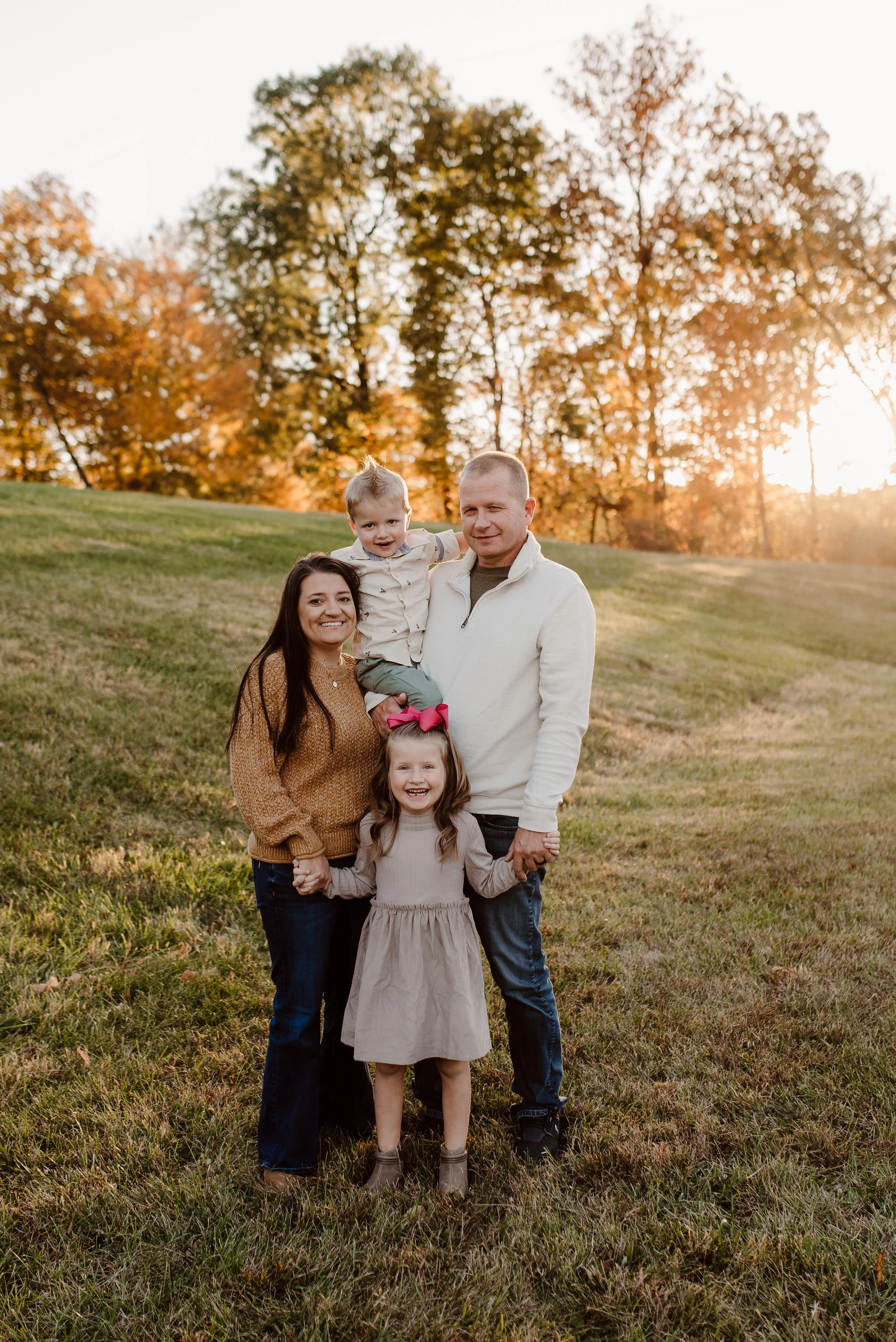 A family of four standing outdoors on a grassy hill during fall, with trees with orange leaves and the setting sun in the background. The mother and father are holding hands, and the children are smiling, with the girl in front and the boy being held