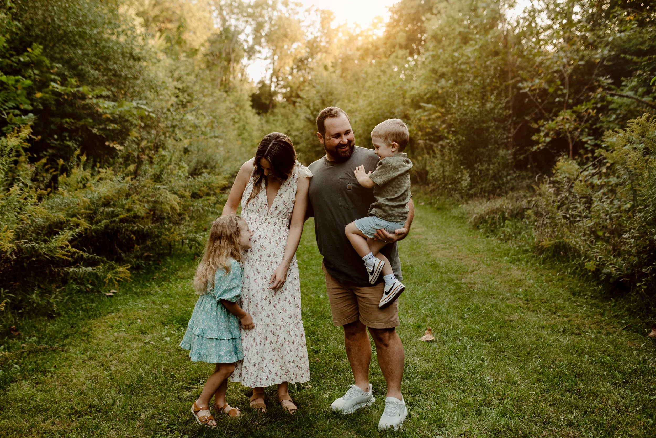A family of four, including a man, woman, and two children, walking and playing in a grassy pathway surrounded by trees in a park during sunset.