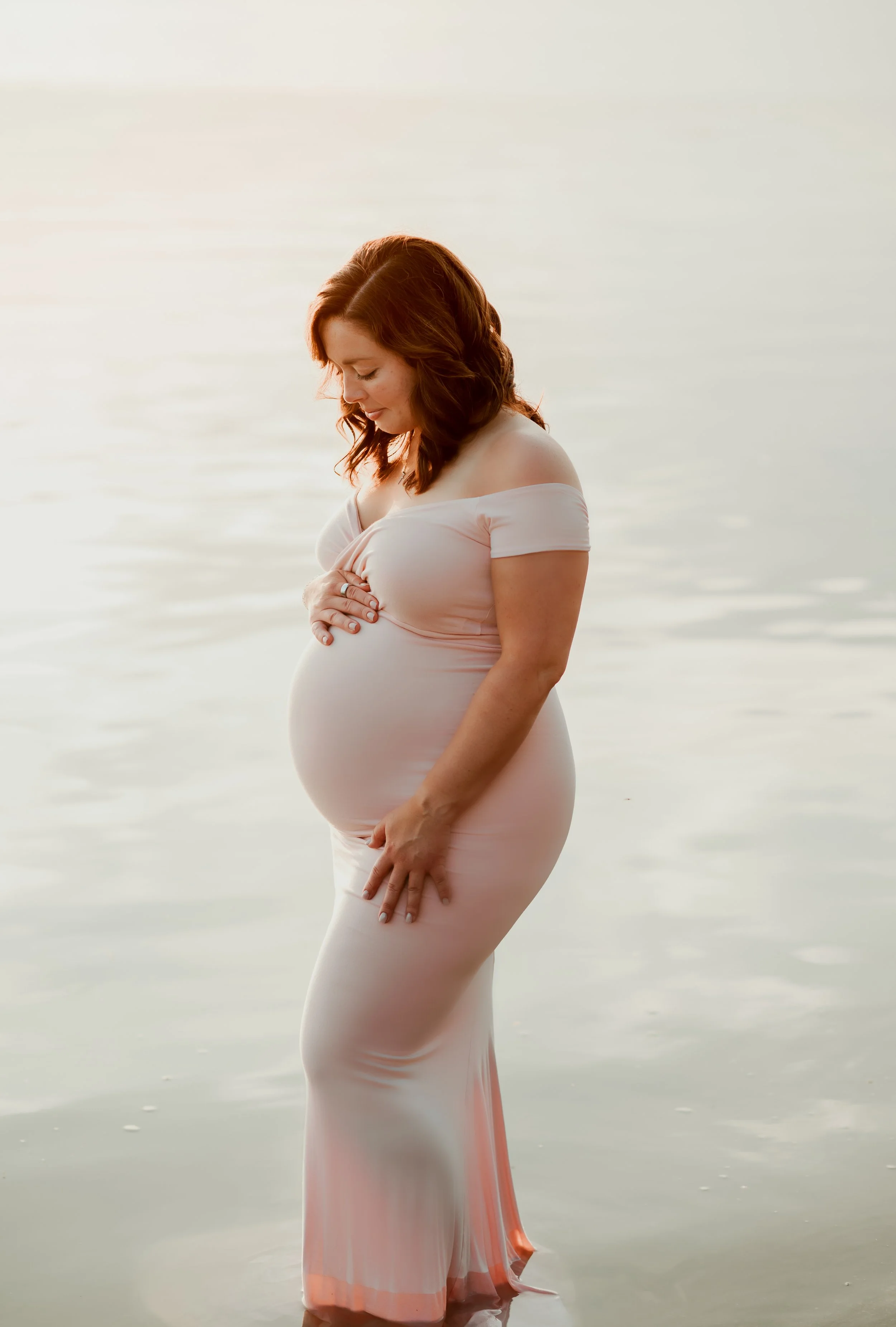 A pregnant woman in a pink, off-shoulder dress standing in shallow water at sunset, looking down with her hand on her belly.