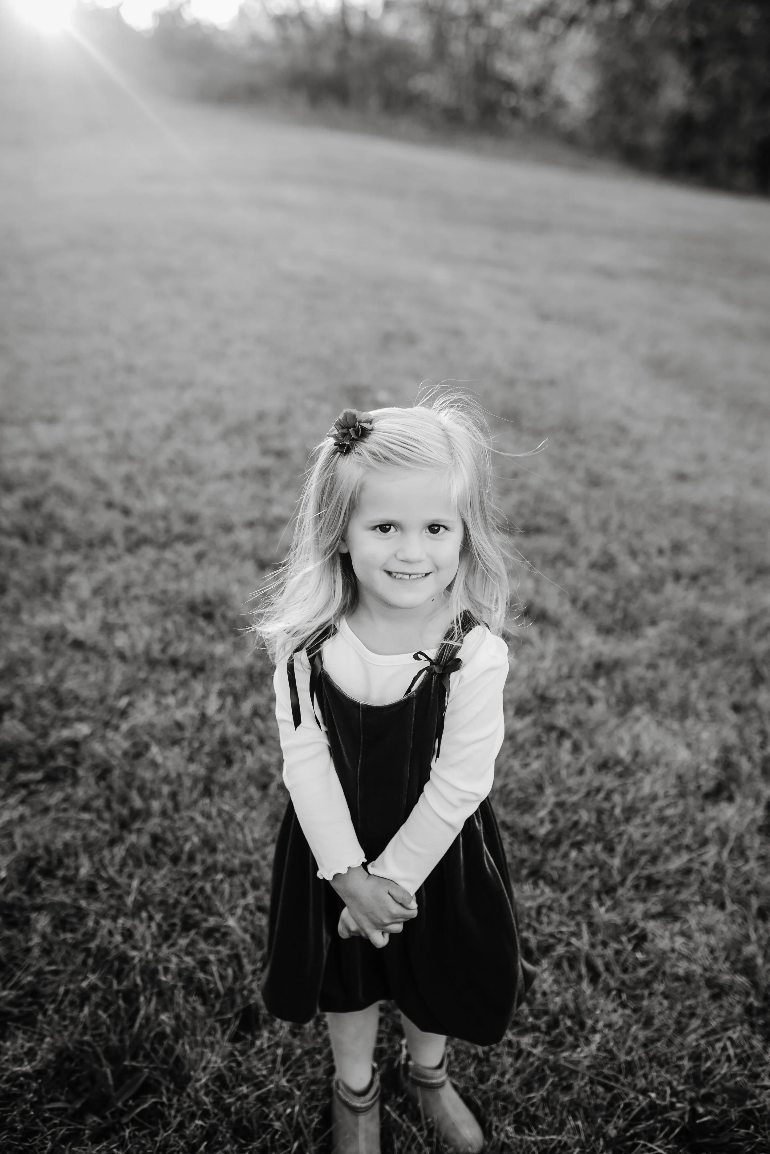 A smiling young girl with light hair standing on a grassy field at sunset, wearing a long-sleeved shirt, a dress, and boots.