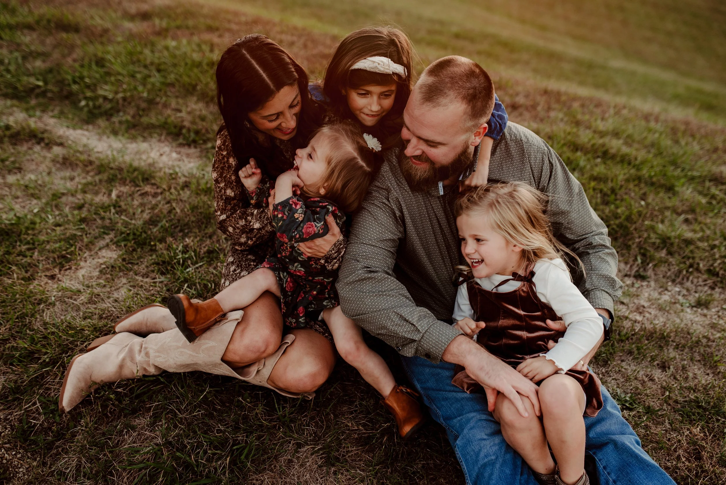 A family of seven, including two adults and five children, sitting on the grass and laughing together outdoors during sunset.