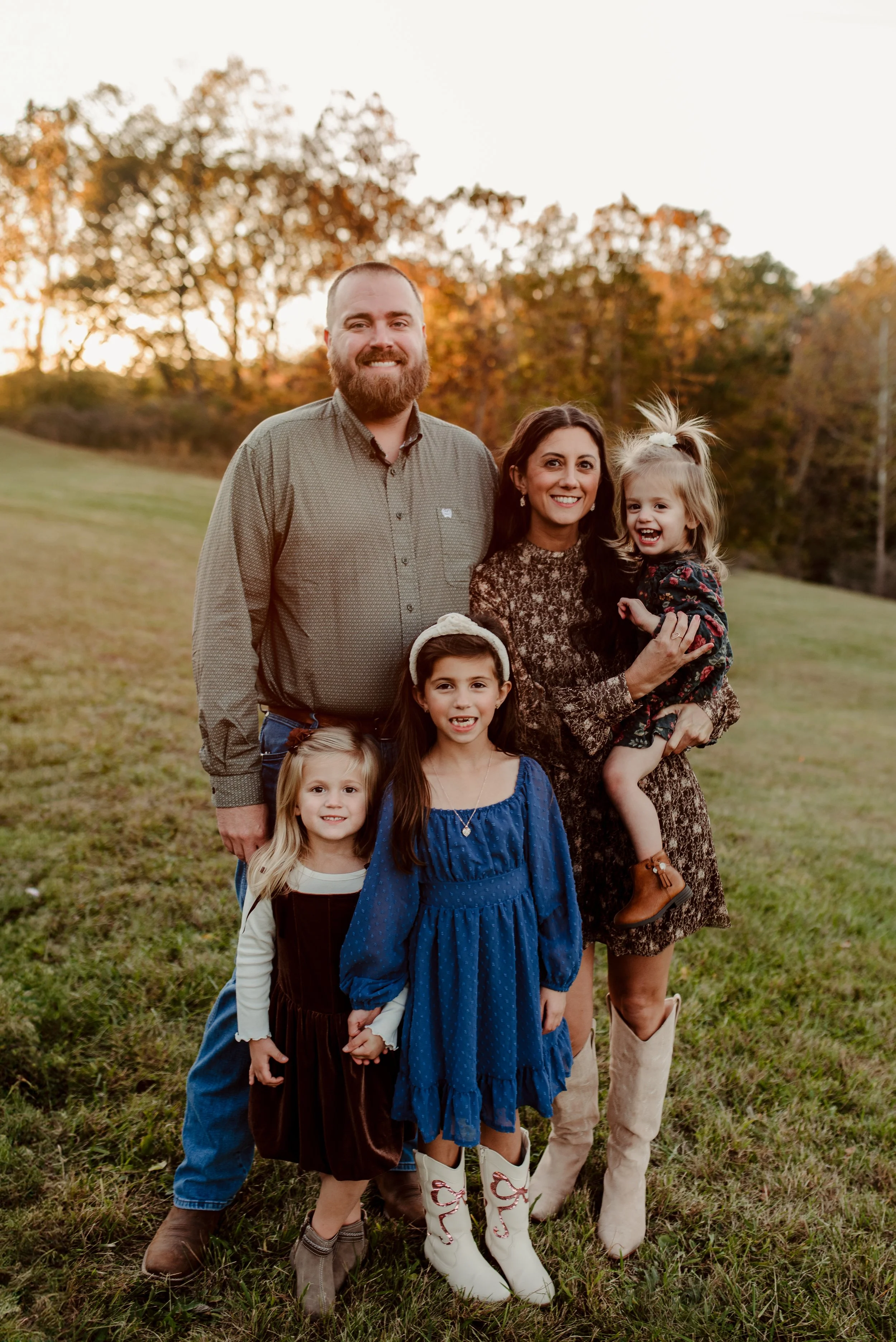 Family photo outdoors during fall, including a man, woman, and four young girls, with trees and a grassy field in the background.