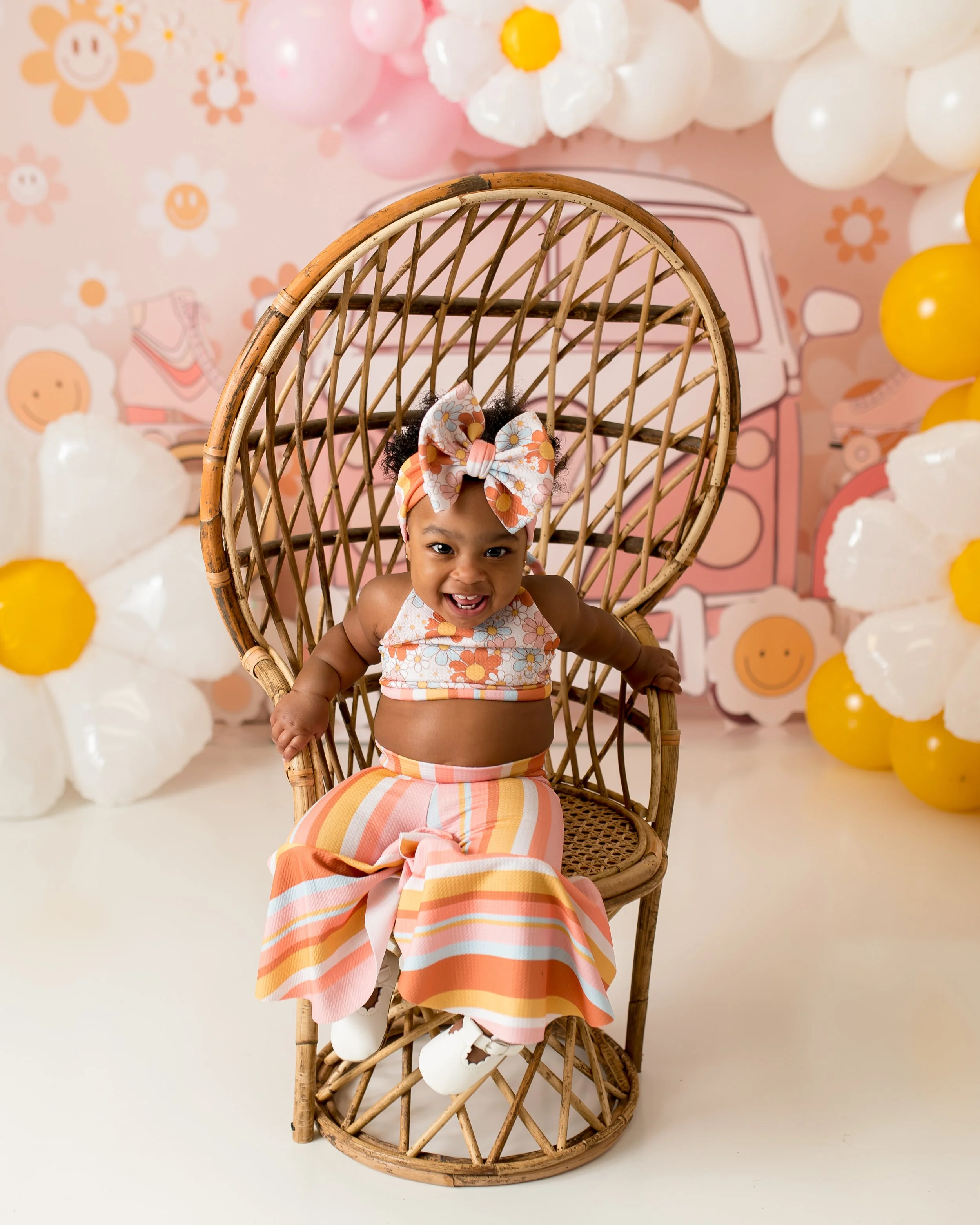 A smiling baby girl sitting on a rattan chair in front of a pastel pink flower-themed backdrop with balloons.