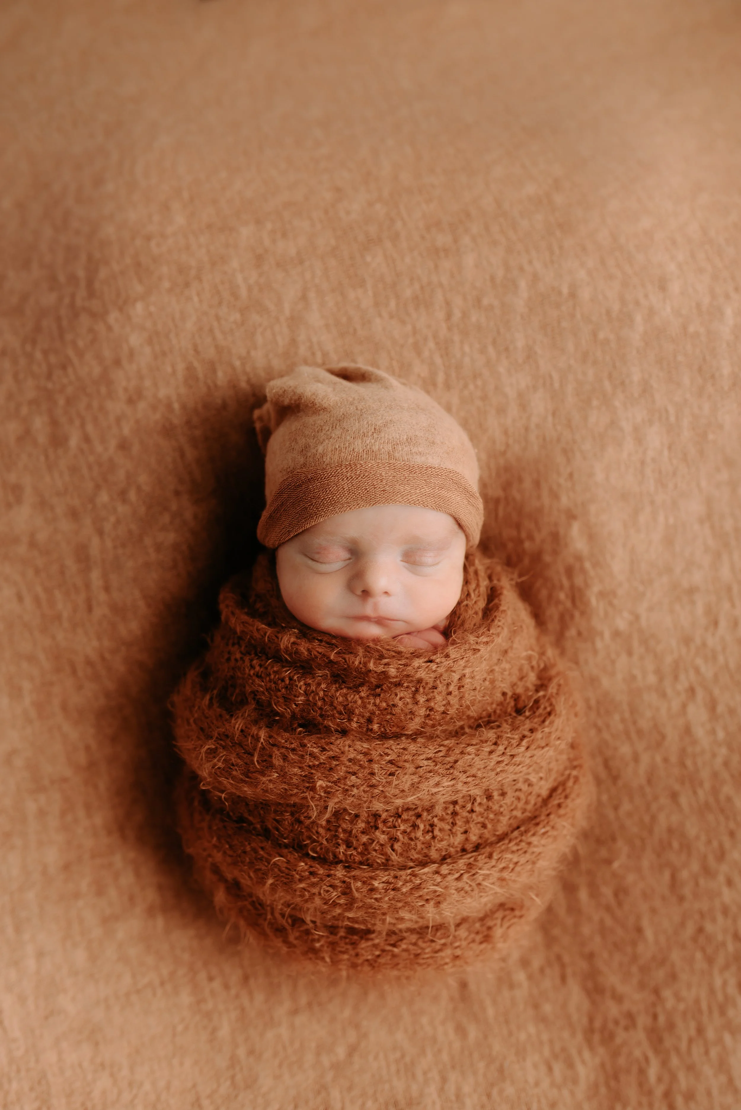 A newborn baby wrapped in a brown knit blanket, wearing a matching brown beanie, sleeping peacefully on a brown textured surface.