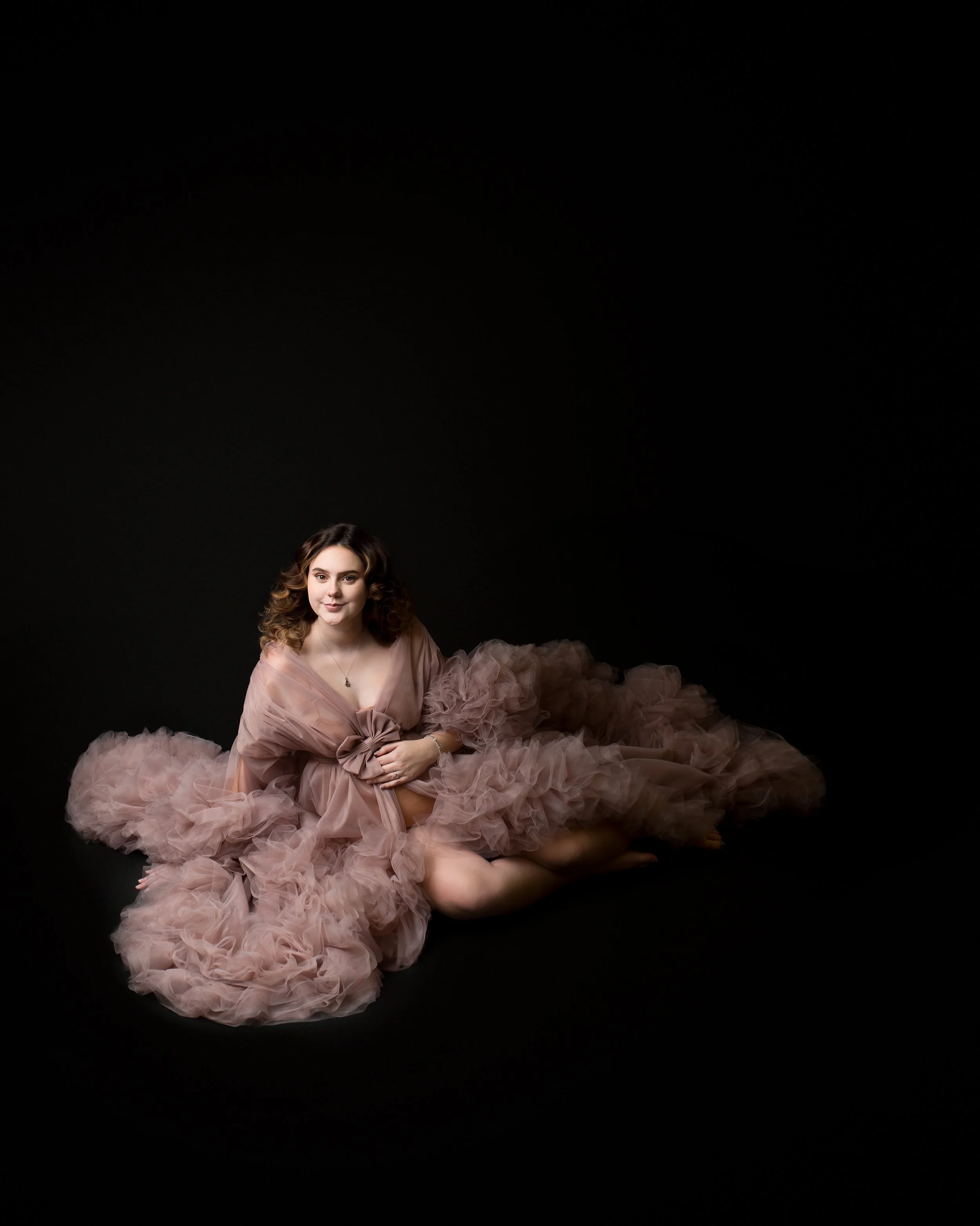 A woman sitting on the floor in a flowing, ruffled pink gown against a black background.
