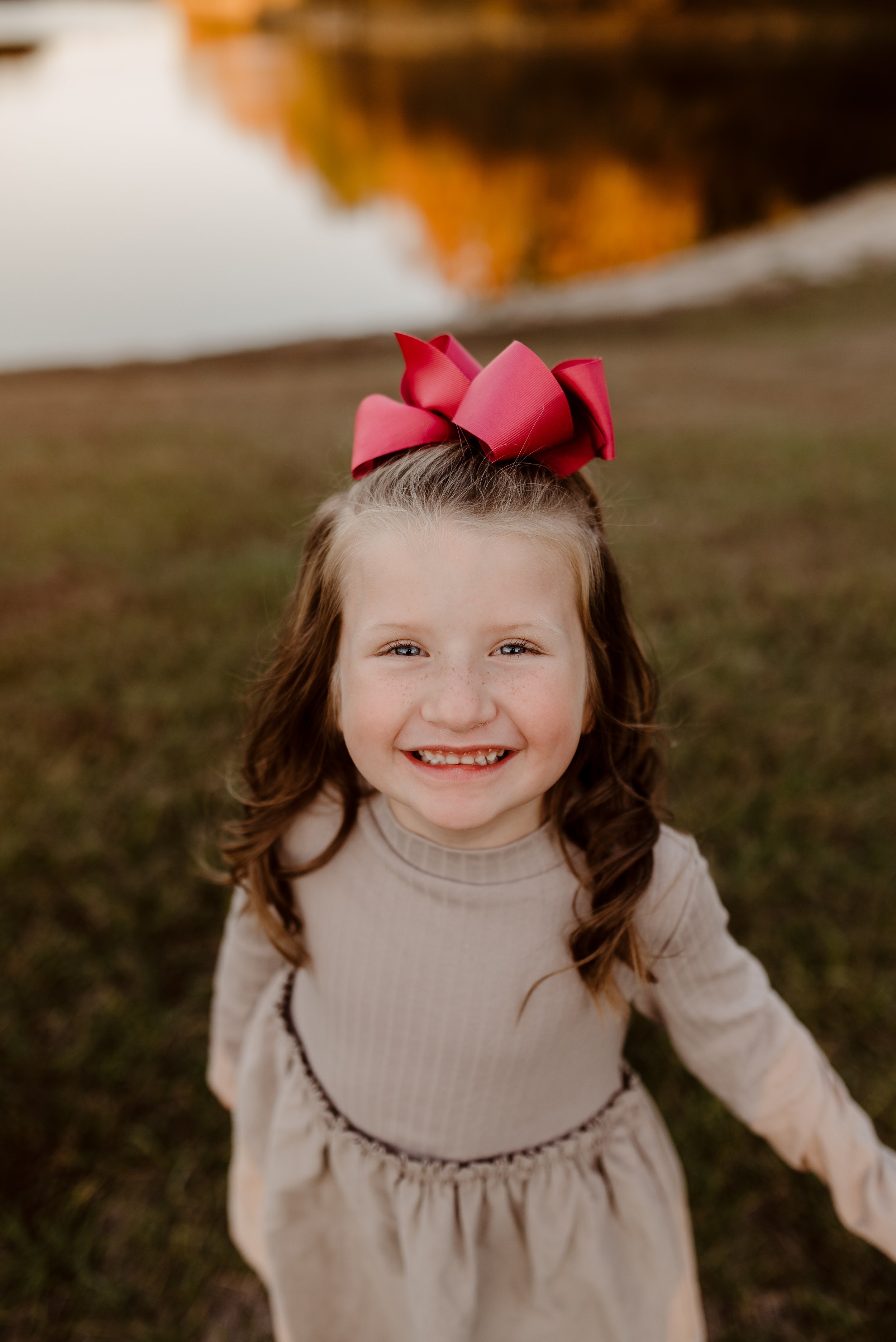 A young girl with long wavy brown hair and a large red bow on her head, smiling outdoors near a body of water during autumn.